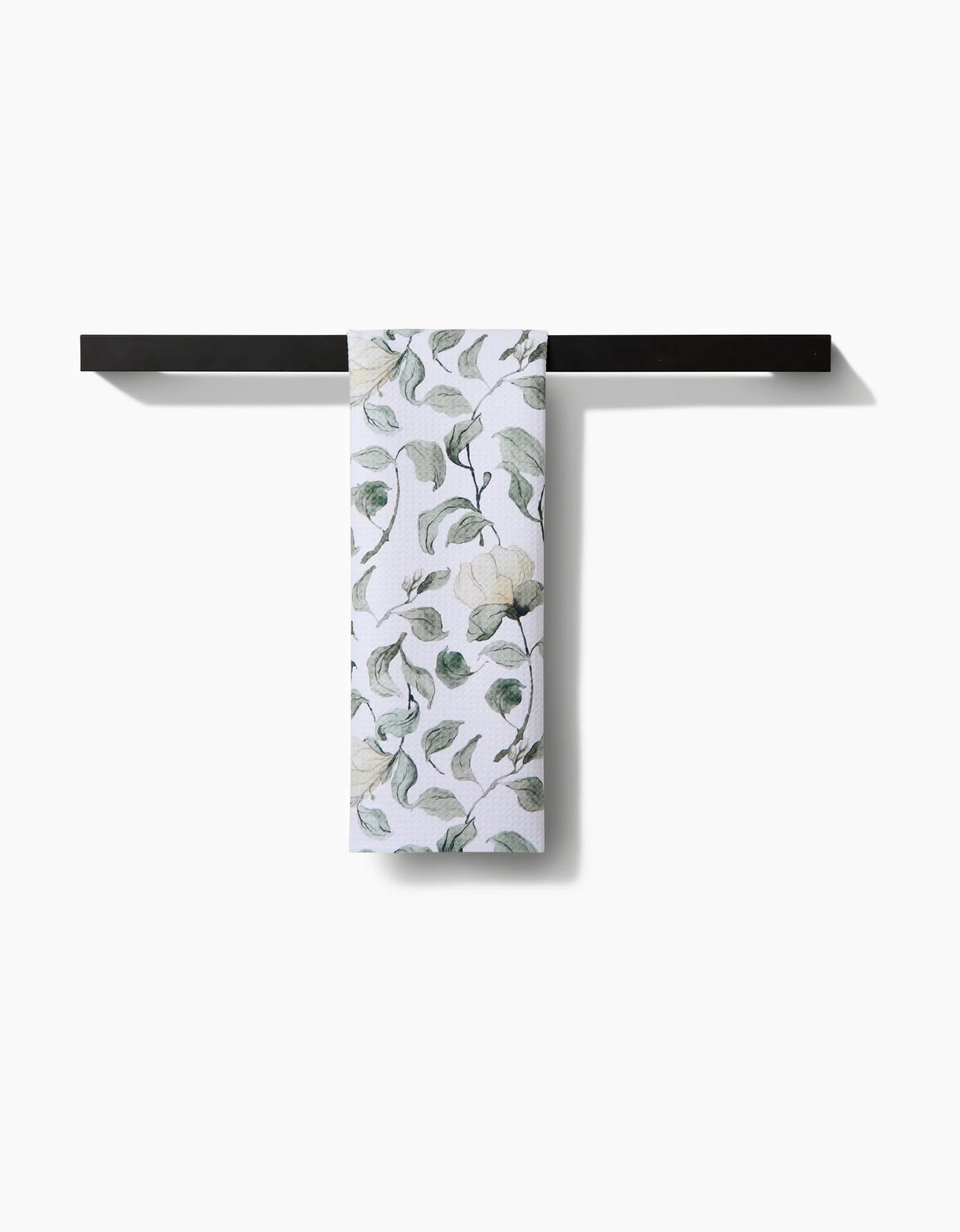 The Aubrey Fairchild Spring Floral Blossoms tea towel, made from recycled materials, features a green and gray leafy floral pattern and hangs over a sleek black towel bar against a plain white background.