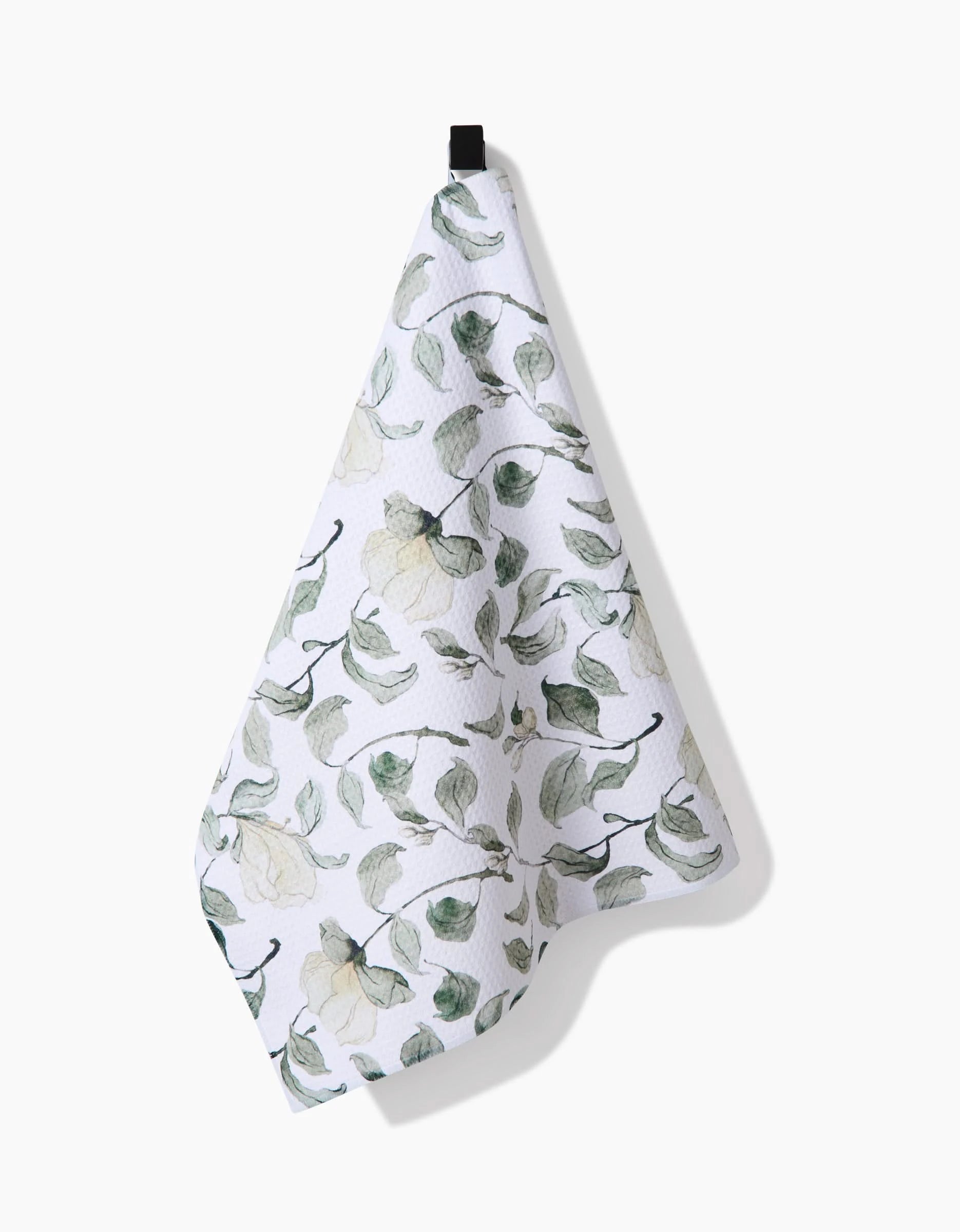 The Spring Floral Blossoms tea towel by Aubrey Fairchild, made from recycled materials in white with a green and yellow leafy fruit pattern, hangs from a black loop against a plain white background.