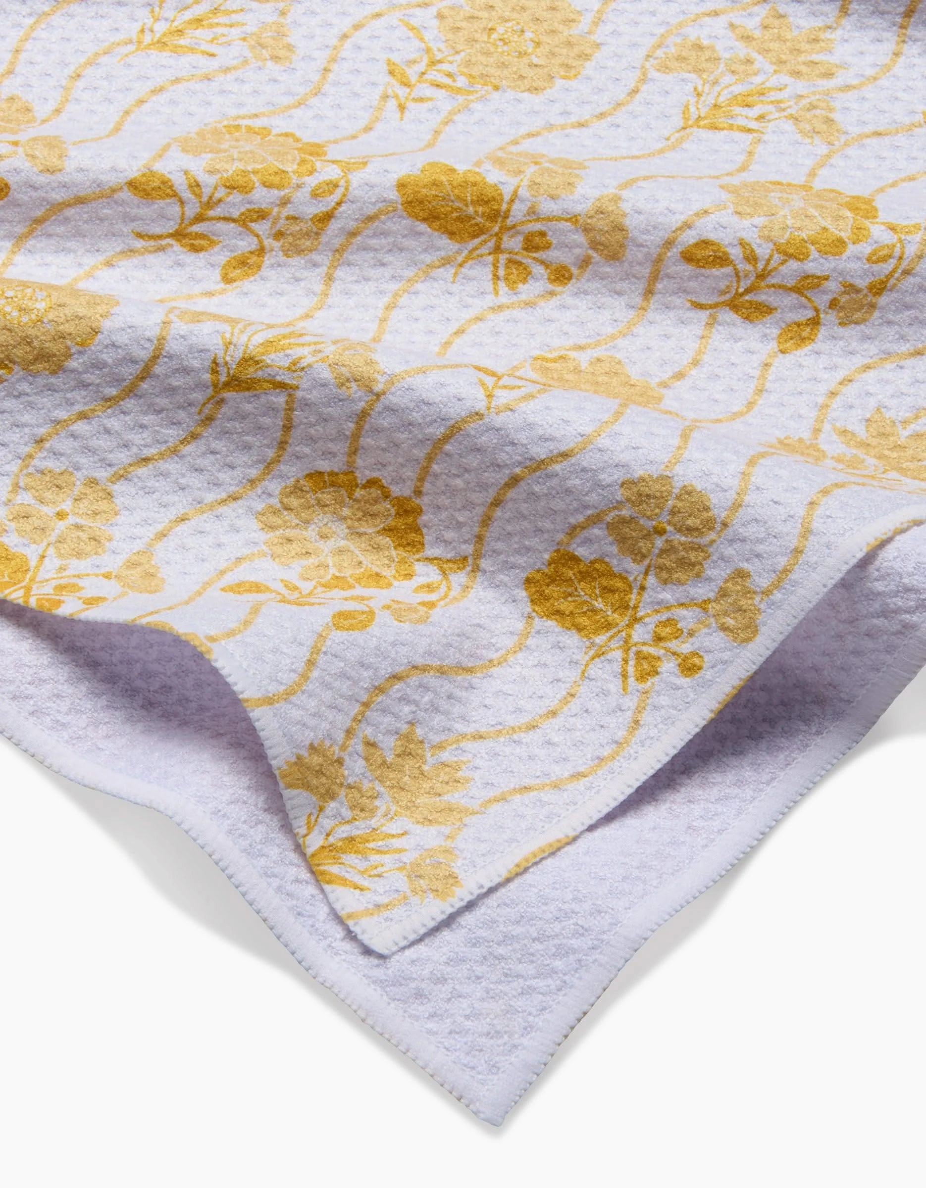 A close-up of the Danelys Sidron Spring Wave tea towel shows white textured Geoweave fabric with a yellow floral and wavy line pattern, one corner folded over. Crafted from recycled materials.