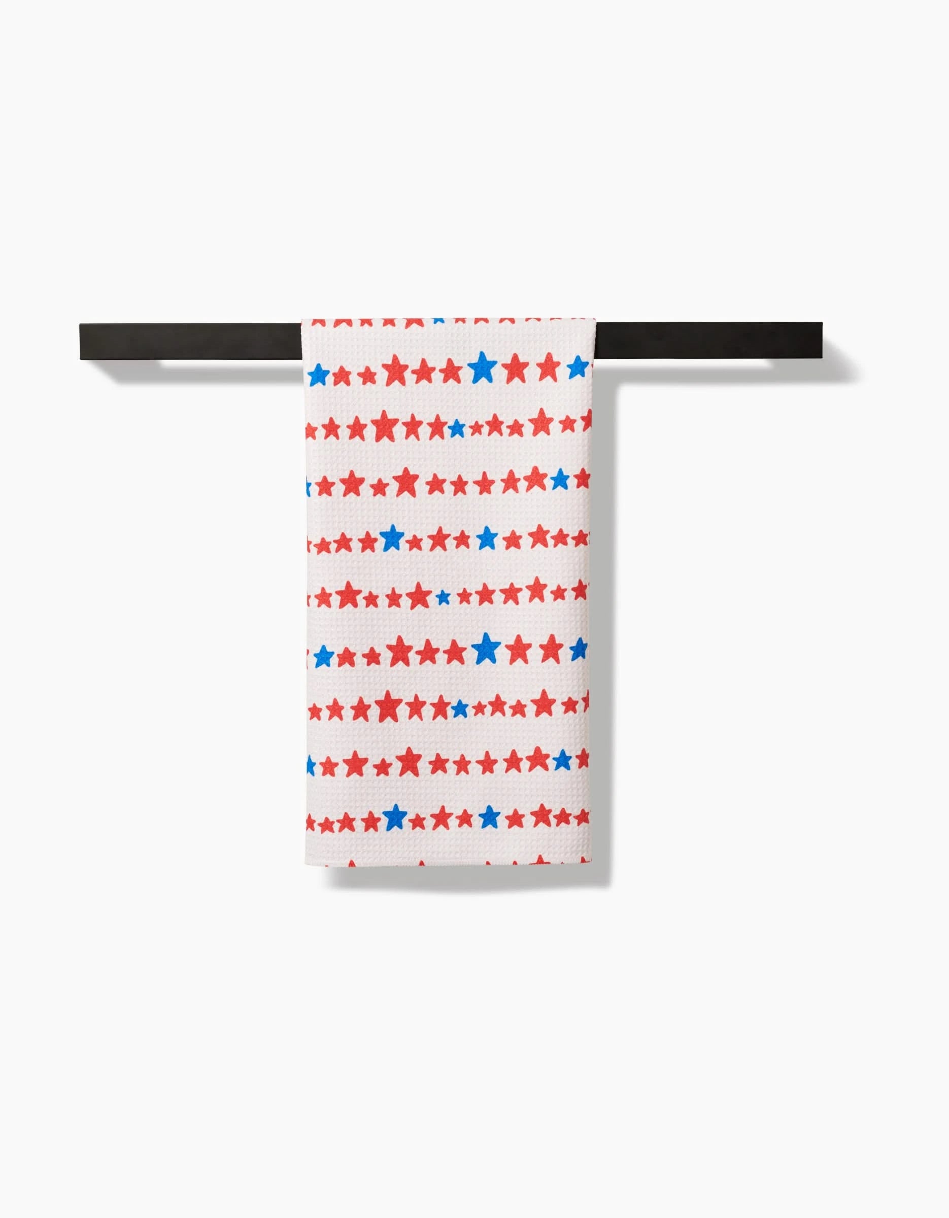 A Kayla Ann Starry Stripes Tea Towel with red and blue stars hangs on a black bar, its Geoweave™ fabric blending style and function against a white background.