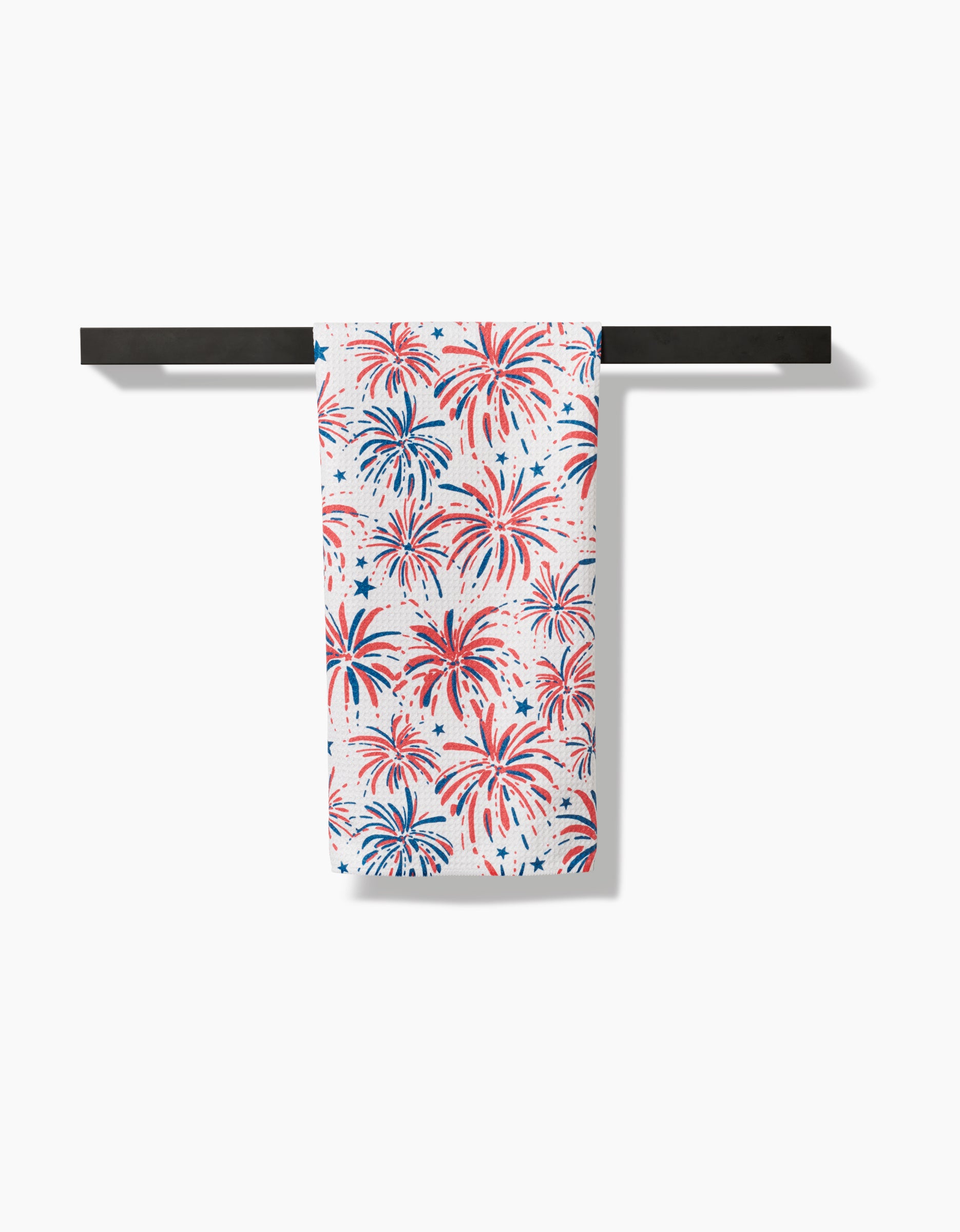 The Stars And Fireworks tea towel by Hufton Studio, featuring a red and blue fireworks pattern on Geoweave fabric, hangs neatly on a black towel bar against a white background.