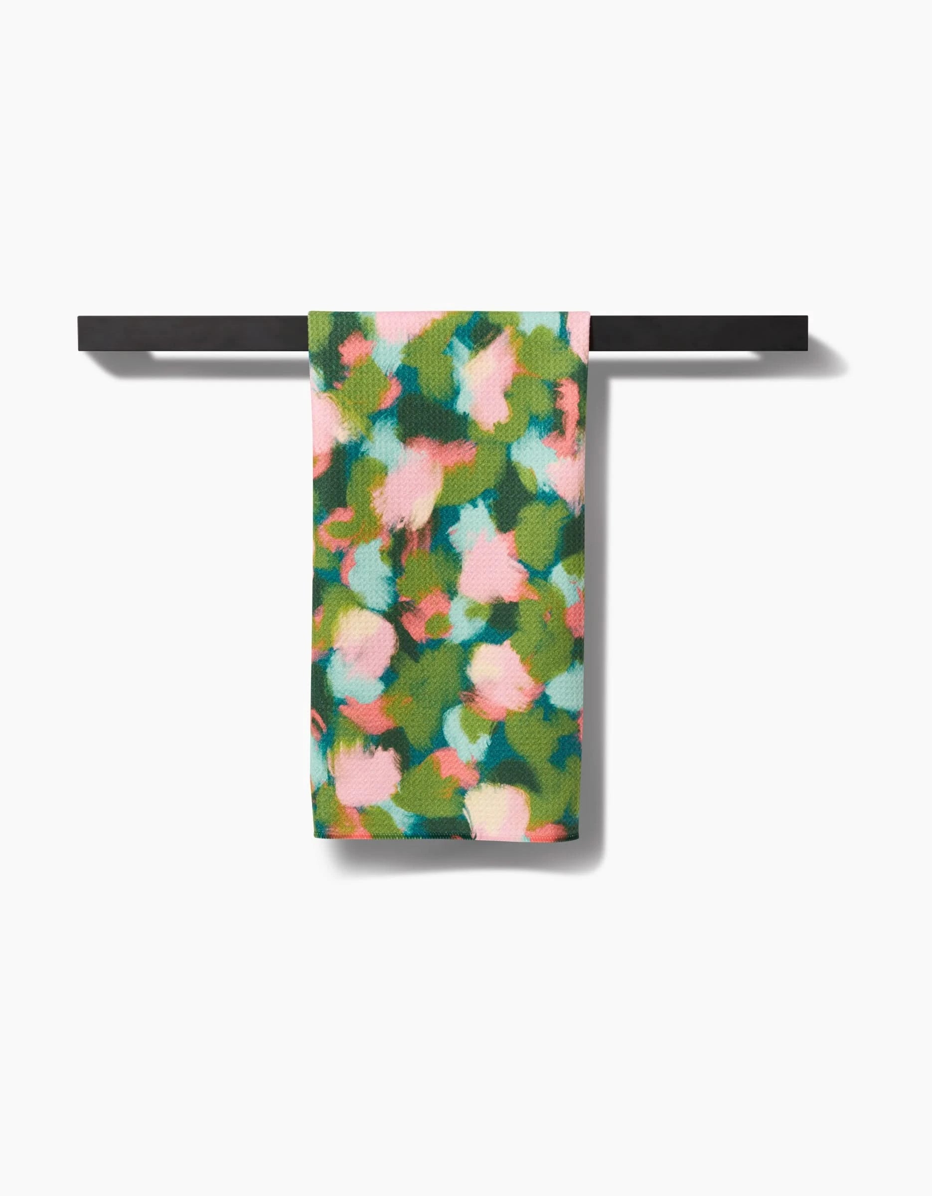 The Stroke of Genius towel by Shelley Smith, featuring a colorful abstract floral design in green, pink, and white on ultra-absorbent Geoweave fabric, hangs neatly over a black towel bar against a white background.