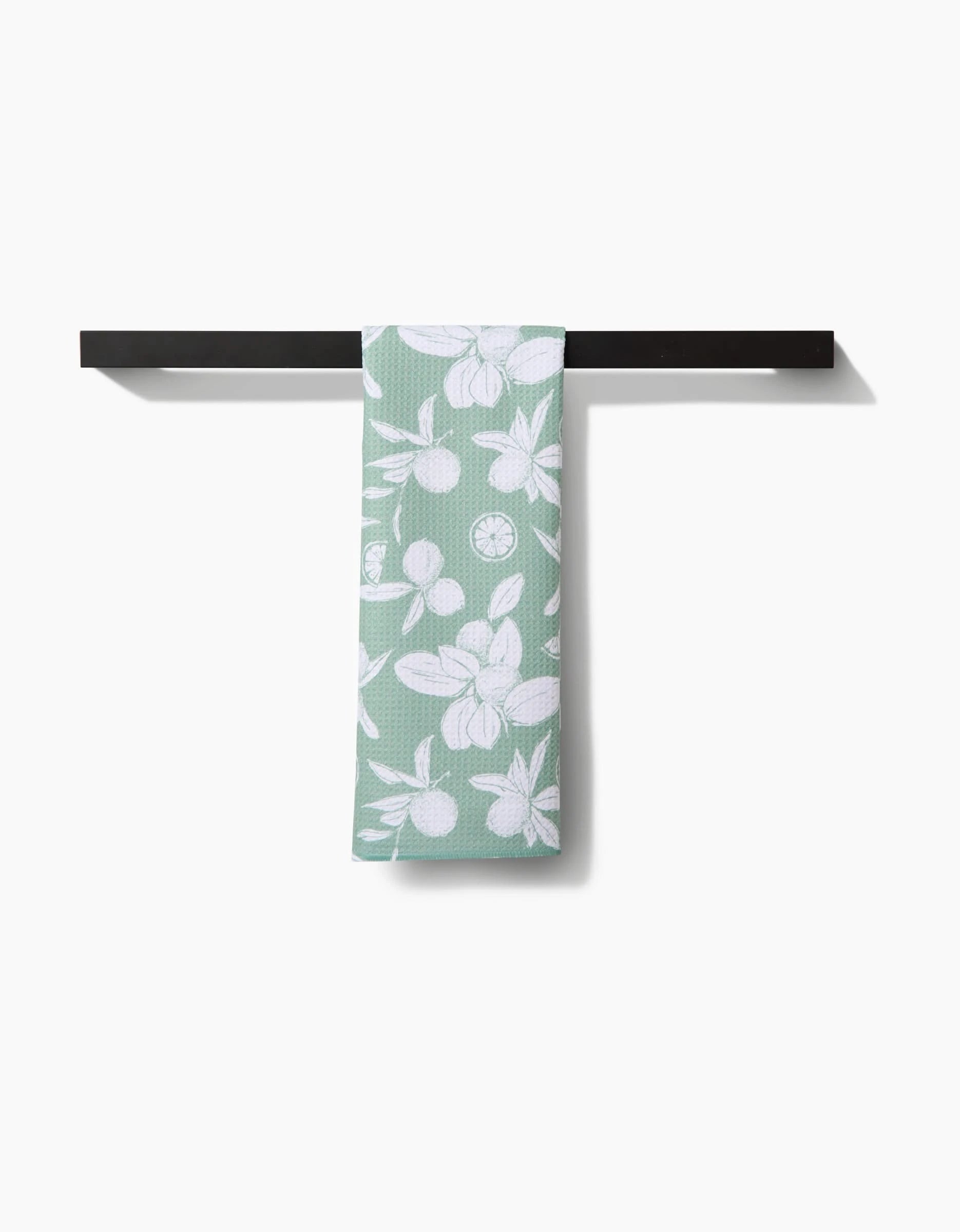 The Hufton Studio Summer Limes tea towel in green, made from durable Geoweave fabric and featuring a white fruit and leaf pattern, is shown hanging neatly on a black towel bar against a white background.