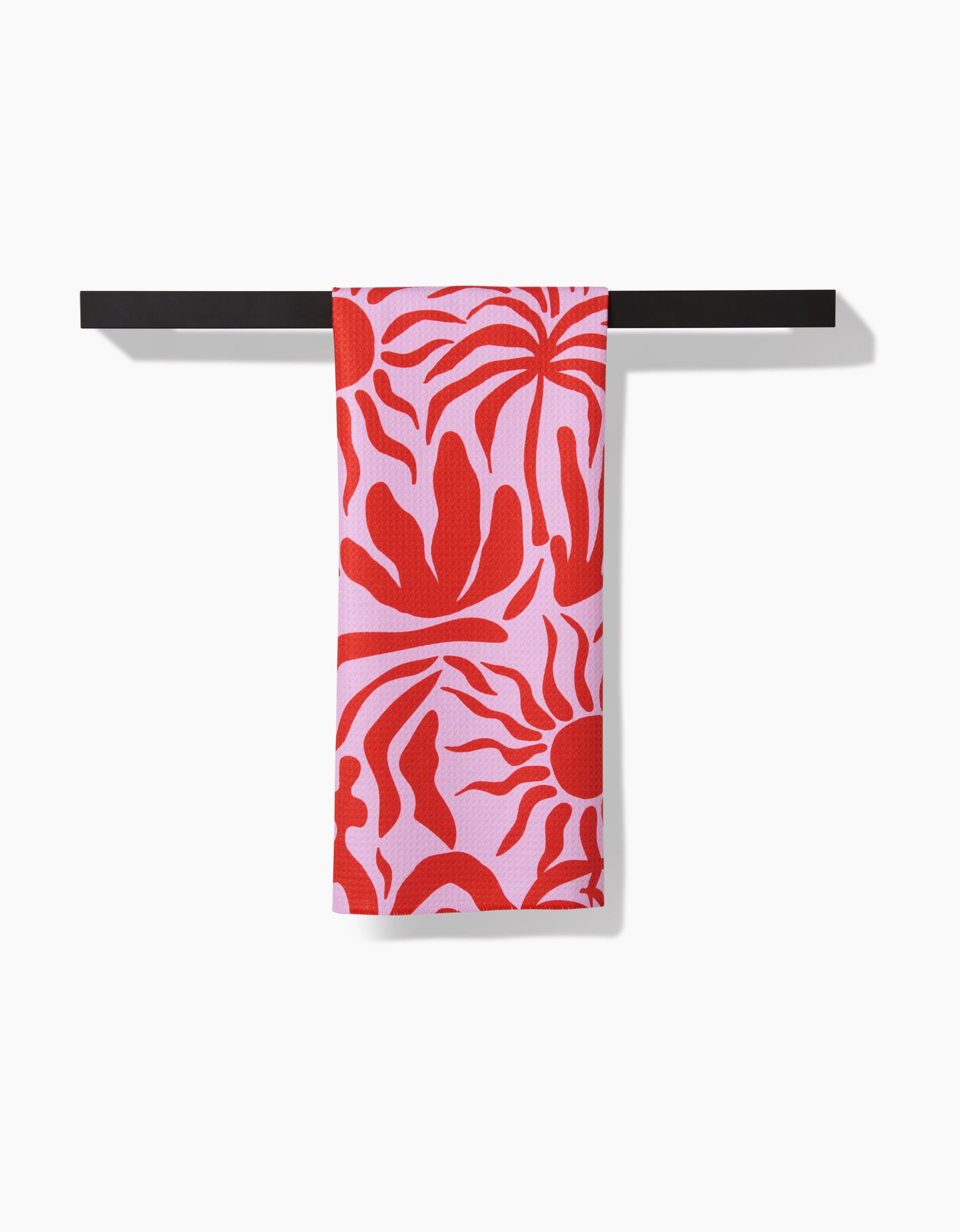 The Summer Sunset Geoweave™ tea towel by Marina Solodka, featuring bold red abstract shapes and sun motifs on pink, hangs neatly over a black bar against a white background.