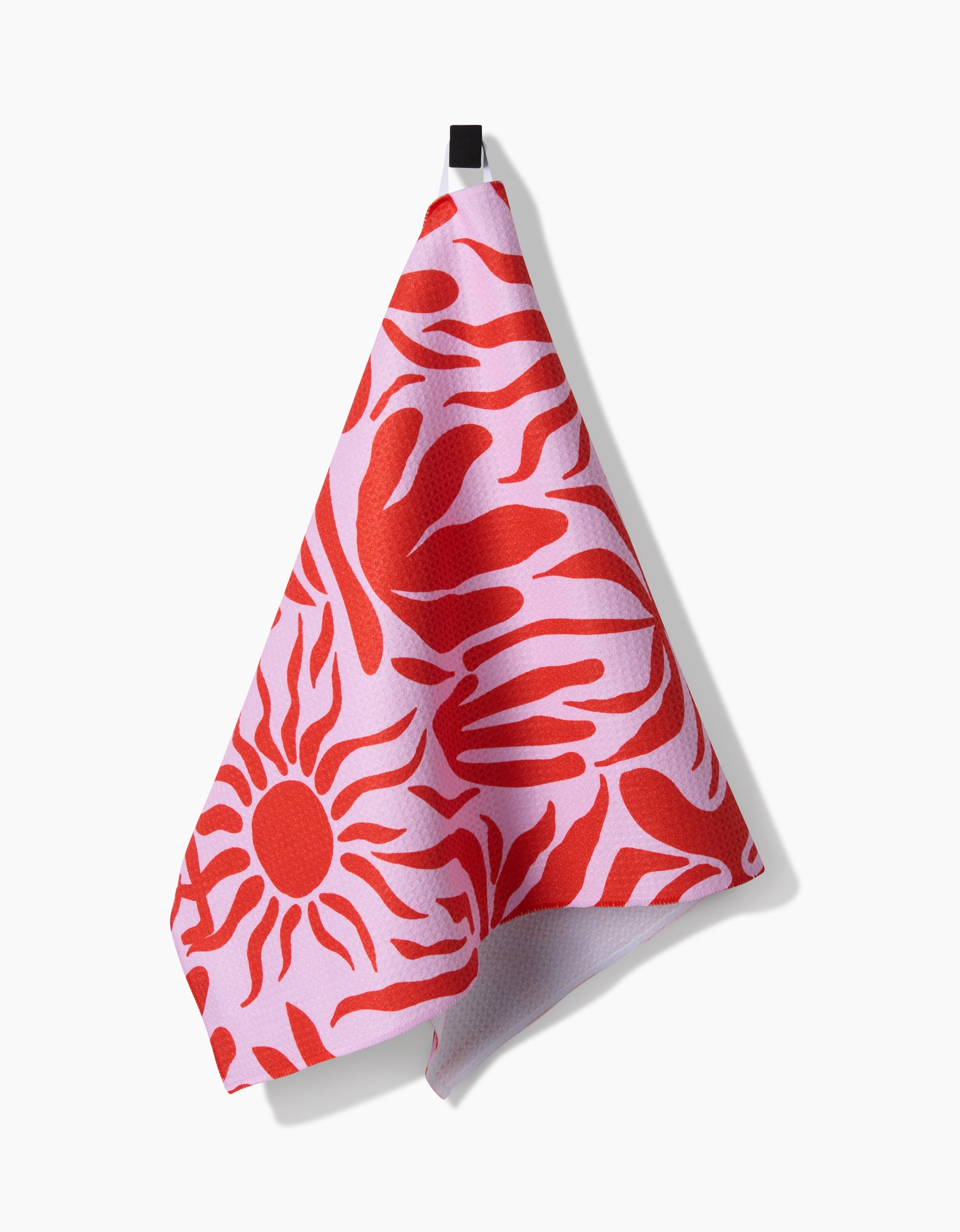 The Summer Sunset tea towel by Marina Solodka features bold red sun and abstract hand patterns on pink, crafted from recycled materials, and hangs from a black loop against a white background.