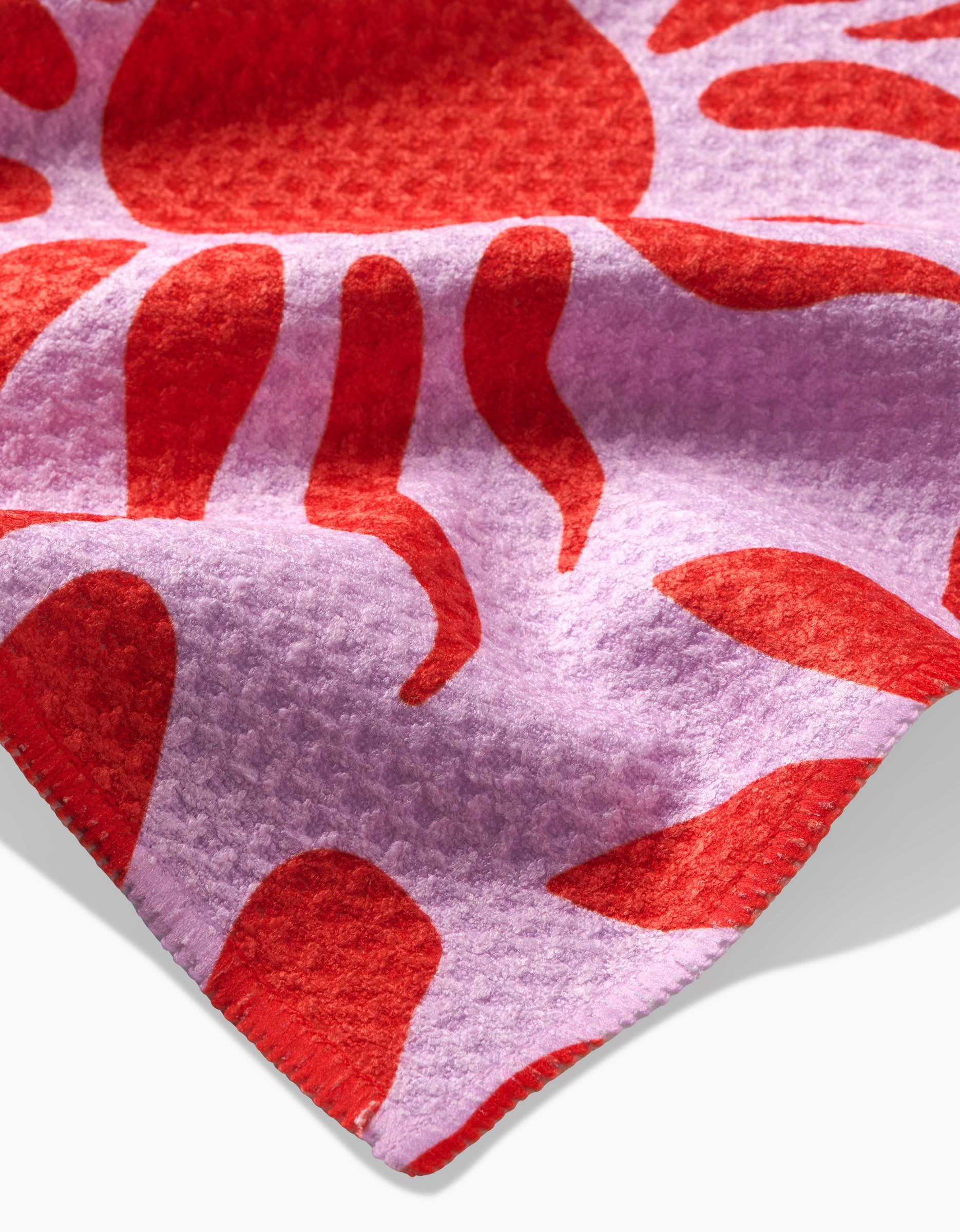 A close-up of the Summer Sunset tea towel by Marina Solodka, featuring textured Geoweave™ fabric with a red sun and wavy rays on a light purple background, highlighting the materials corner.