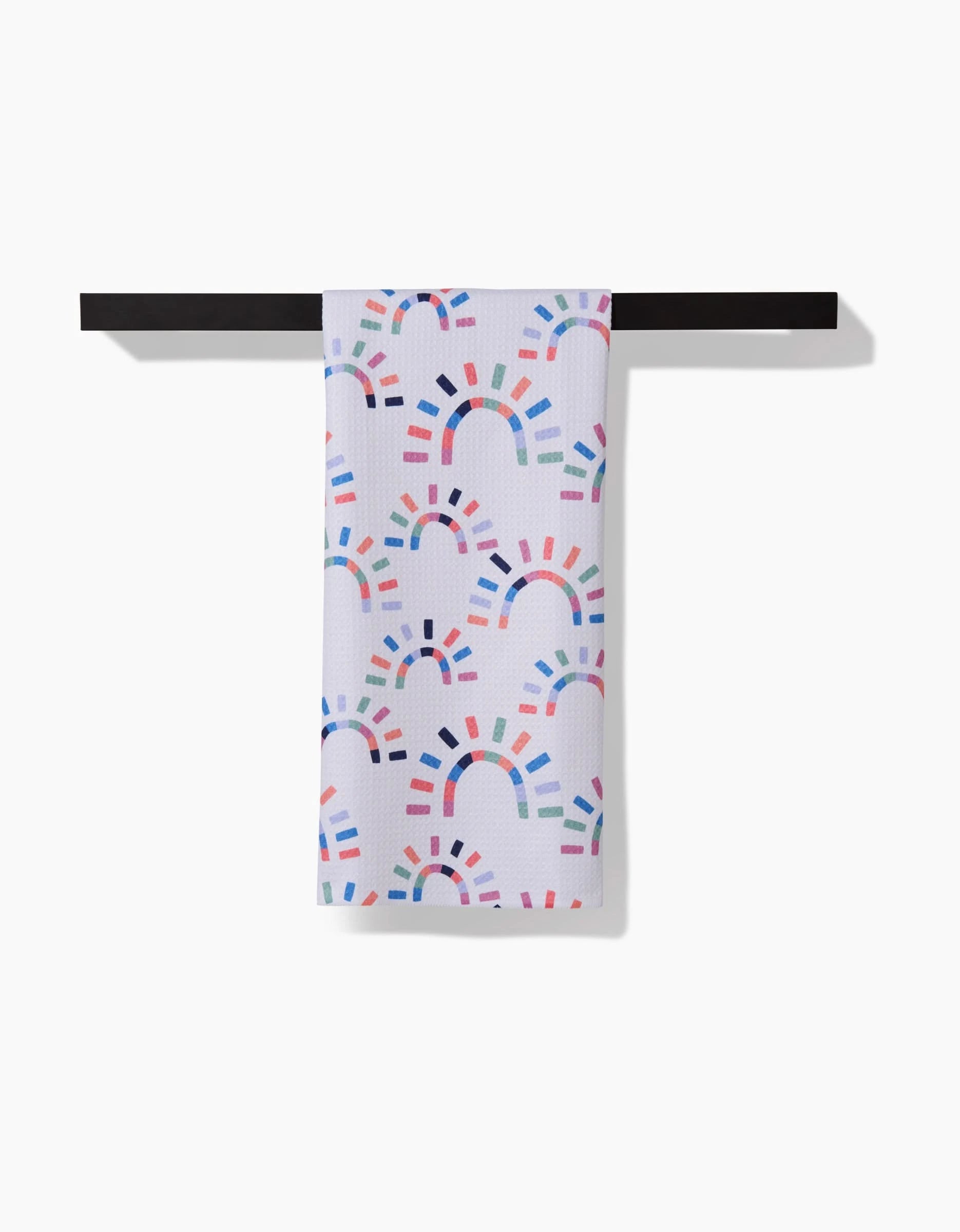 The Sunshine Rainbow towel by Claire Iglesias features rainbow-like arches of multicolored lines on white, hanging over a black rod. Made from ultra-absorbent Geoweave fabric, it stands out beautifully against the white background.