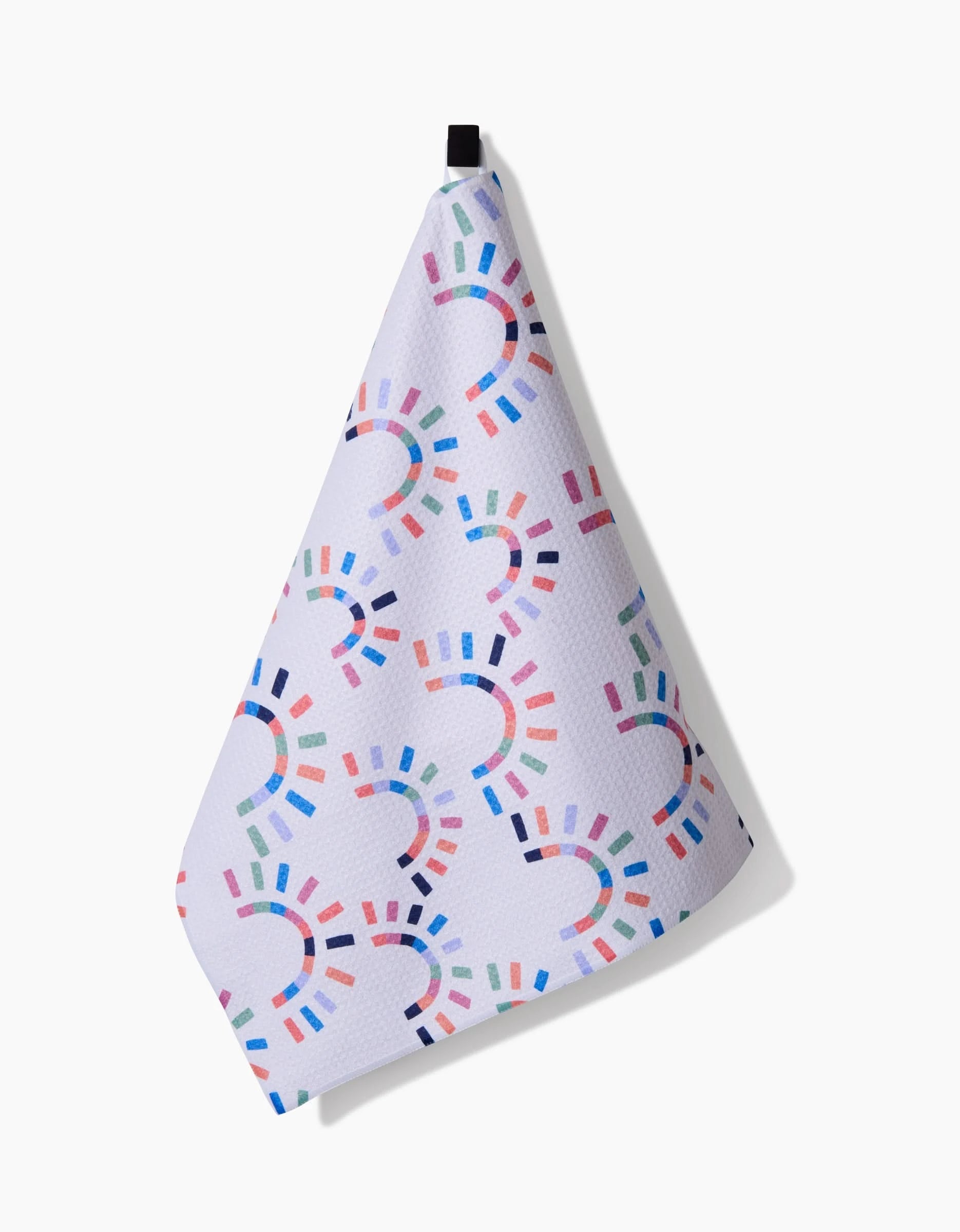 The Sunshine Rainbow towel by Claire Iglesias features a white, ultra-absorbent Geoweave fabric with textured surface and colorful semi-circle patterns in blue, pink, green, and purple. It hangs from a black loop against a white background.