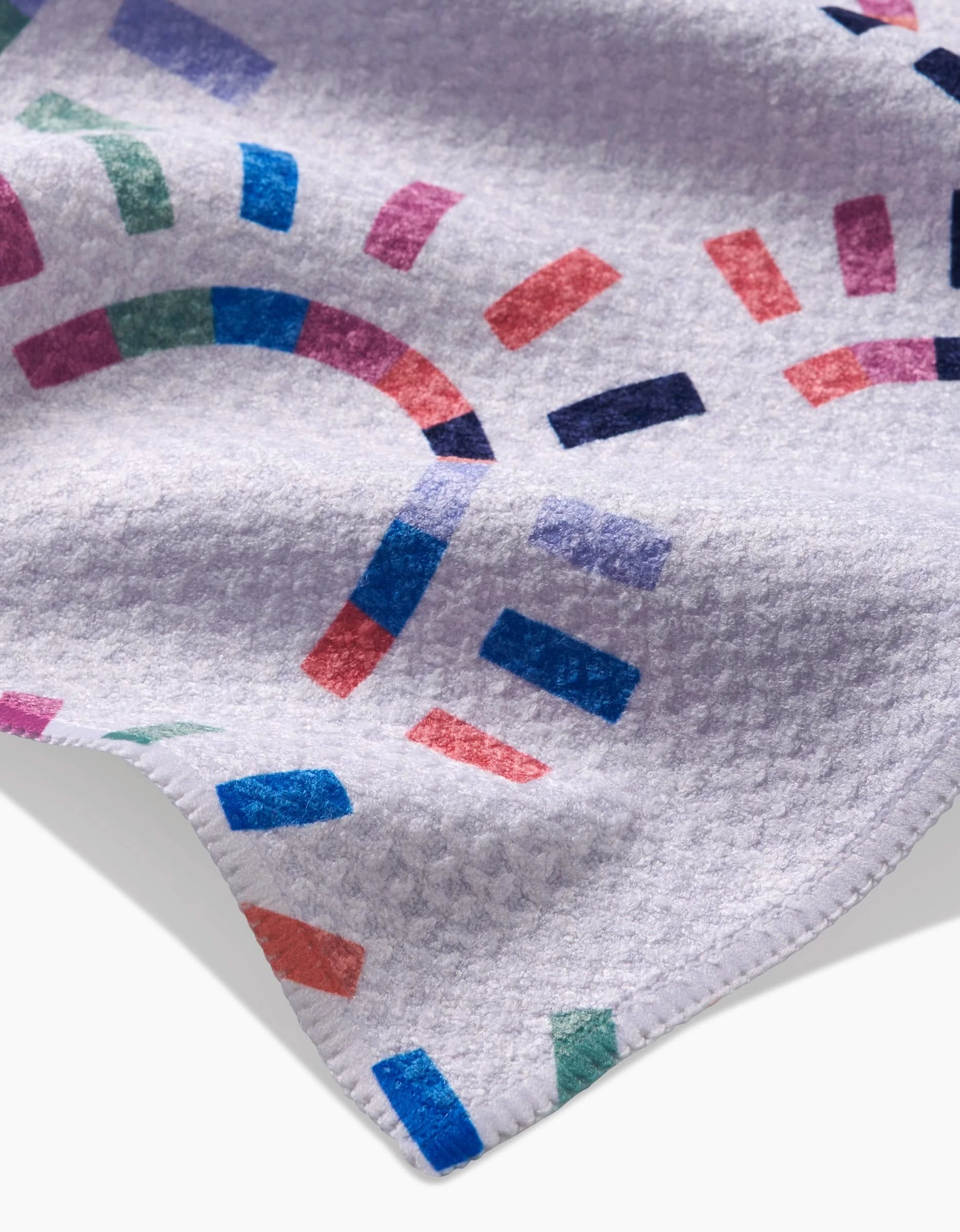 A close-up of Claire Iglesias Sunshine Rainbow towel, made from ultra-absorbent Geoweave fabric with a colorful geometric pattern in blue, purple, green, red, and pink. The white towel features neatly stitched edges.