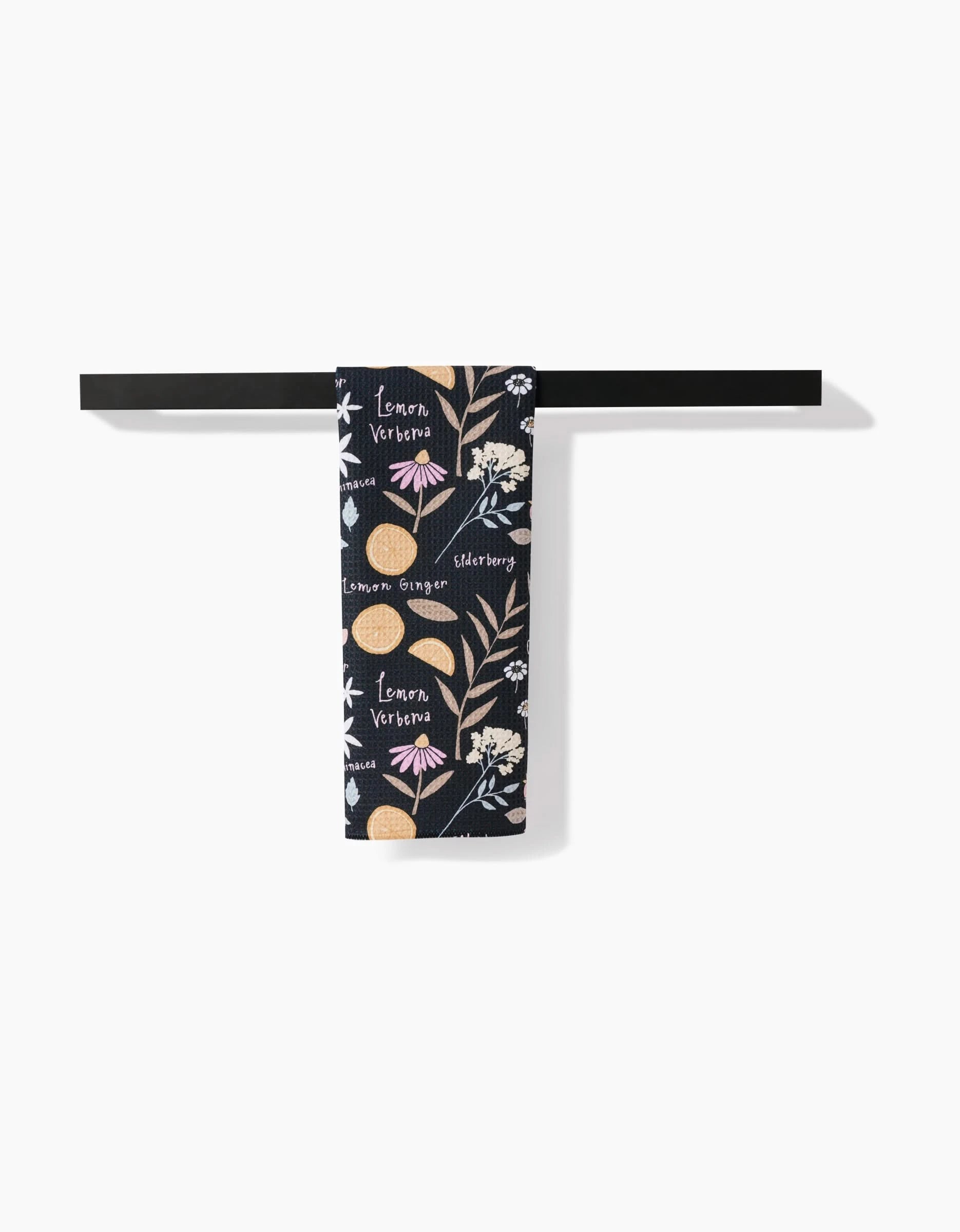 The Sweet Tea Tea Towel by Janna Sue Design features a dark blue floral and lemon pattern with words like Lemon Verbena and elderberry. Made of Geoweave™ fabric, it hangs neatly on a black bar, standing out against a white background.