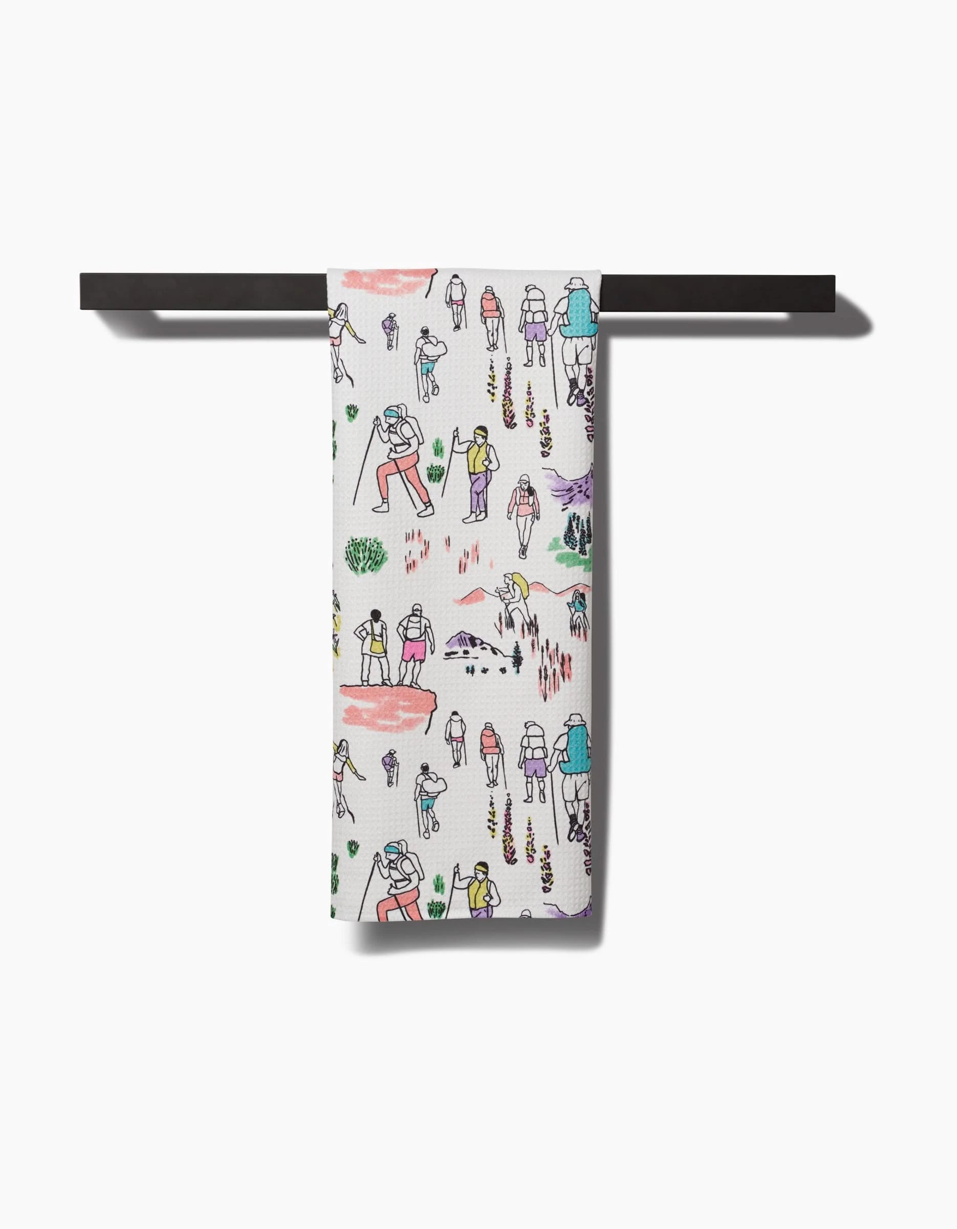 The Take A Hike Tea Towel by Elisabeth Wing features colorful illustrations of people enjoying outdoor adventures, made from recycled materials, and is shown draped over a black bar against a white background.