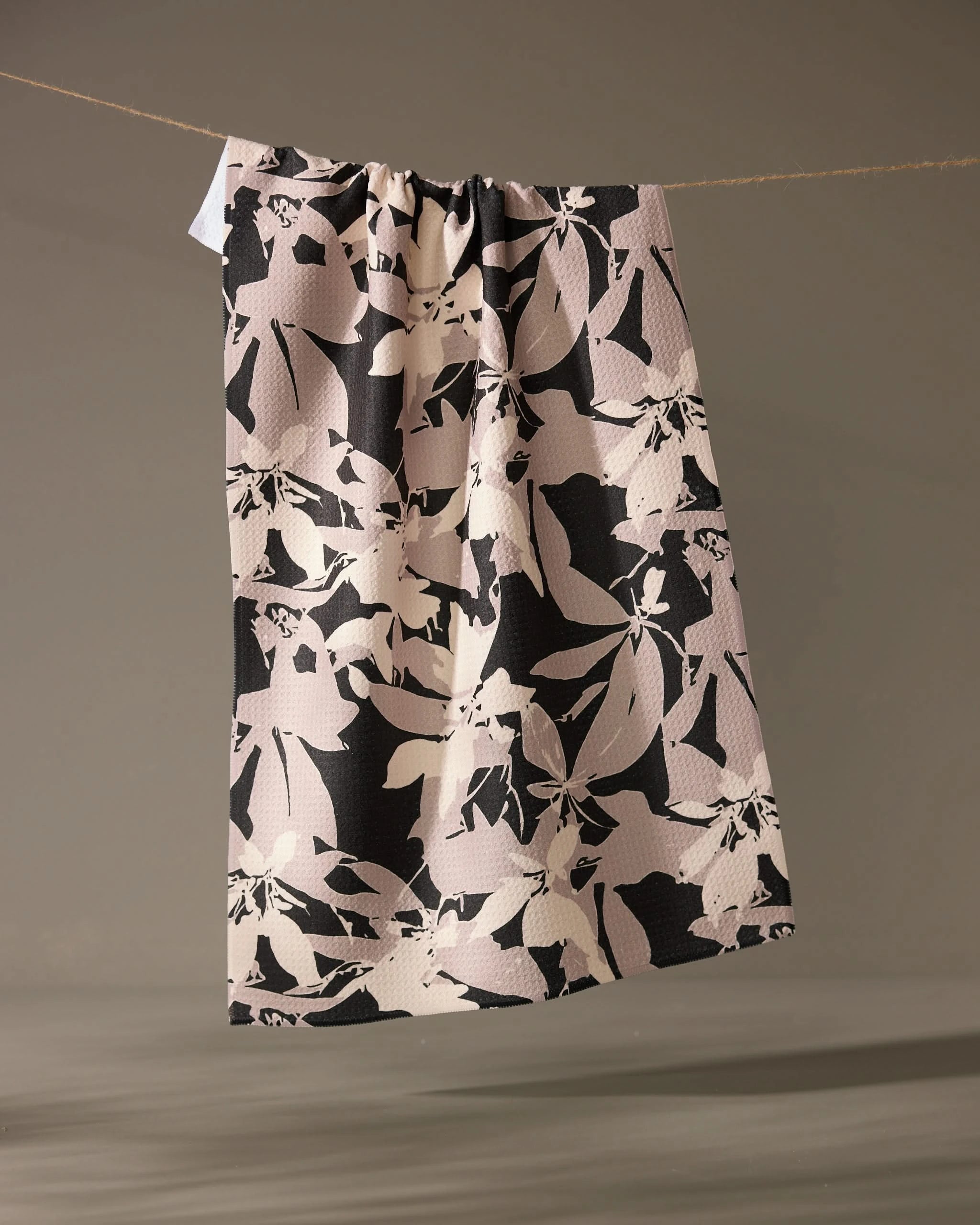 The Tawny tea towel by Geometry, featuring a beige and black abstract floral design, hangs over a thin rope against a neutral background.