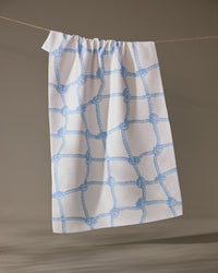 The Geometry Tethered Blue ultra-absorbent dish towel, featuring a blue rope knot pattern on white, hangs from a beige string against a neutral background.