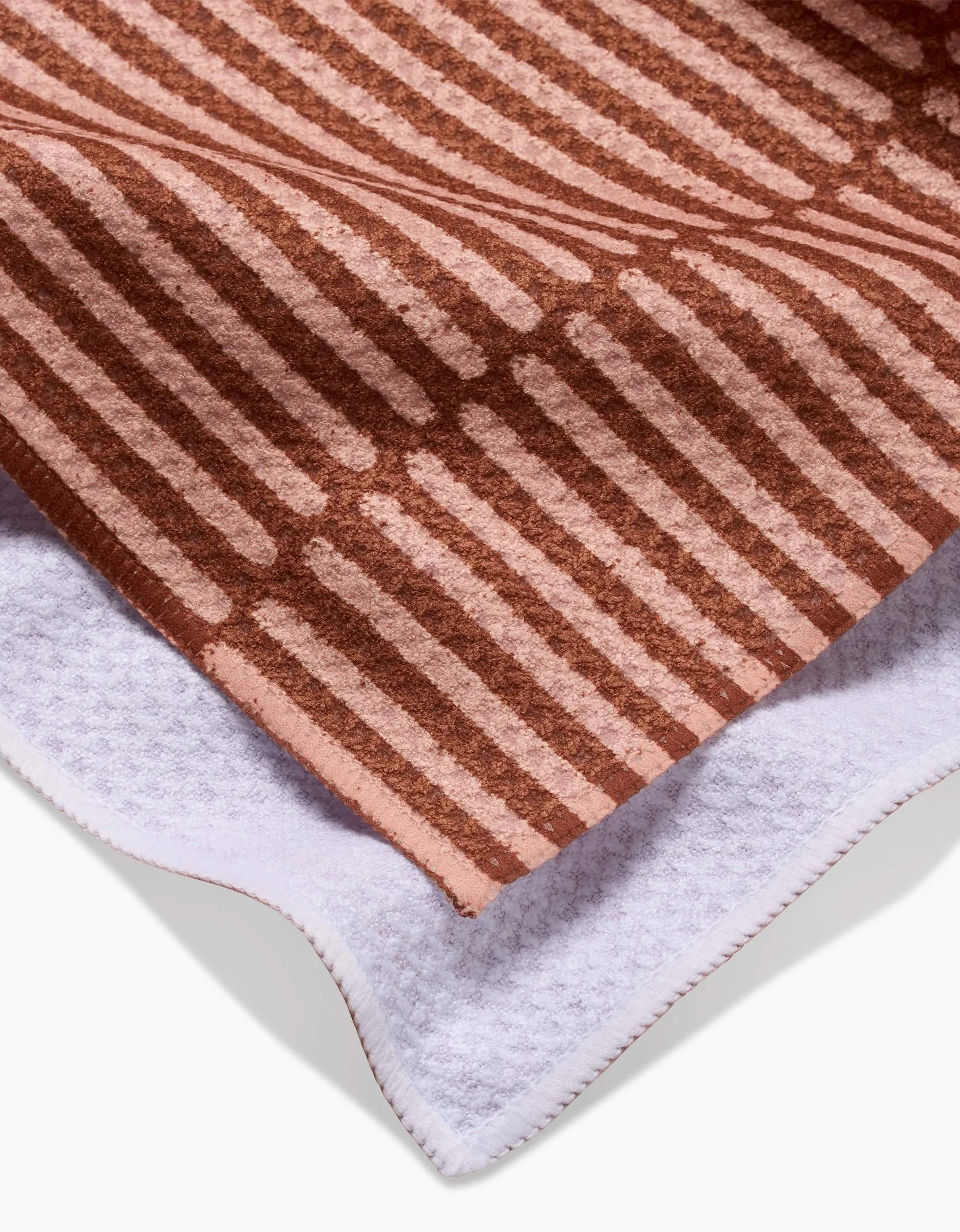 A close-up of a folded Rosa Kichline Textured Stripe Tea Towel shows a brown and pink striped pattern on one side and a plain white ultra-absorbent Geoweave fabric surface on the other.