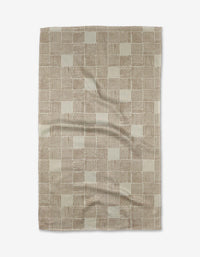 The Thatcher Tea Towel by Studio Jeannot is a rectangular beige and brown towel with a checkered pattern of small squares and stripes, made from ultra-absorbent Geoweave™ fabric for superior performance. Wrinkles are visible when laid flat.