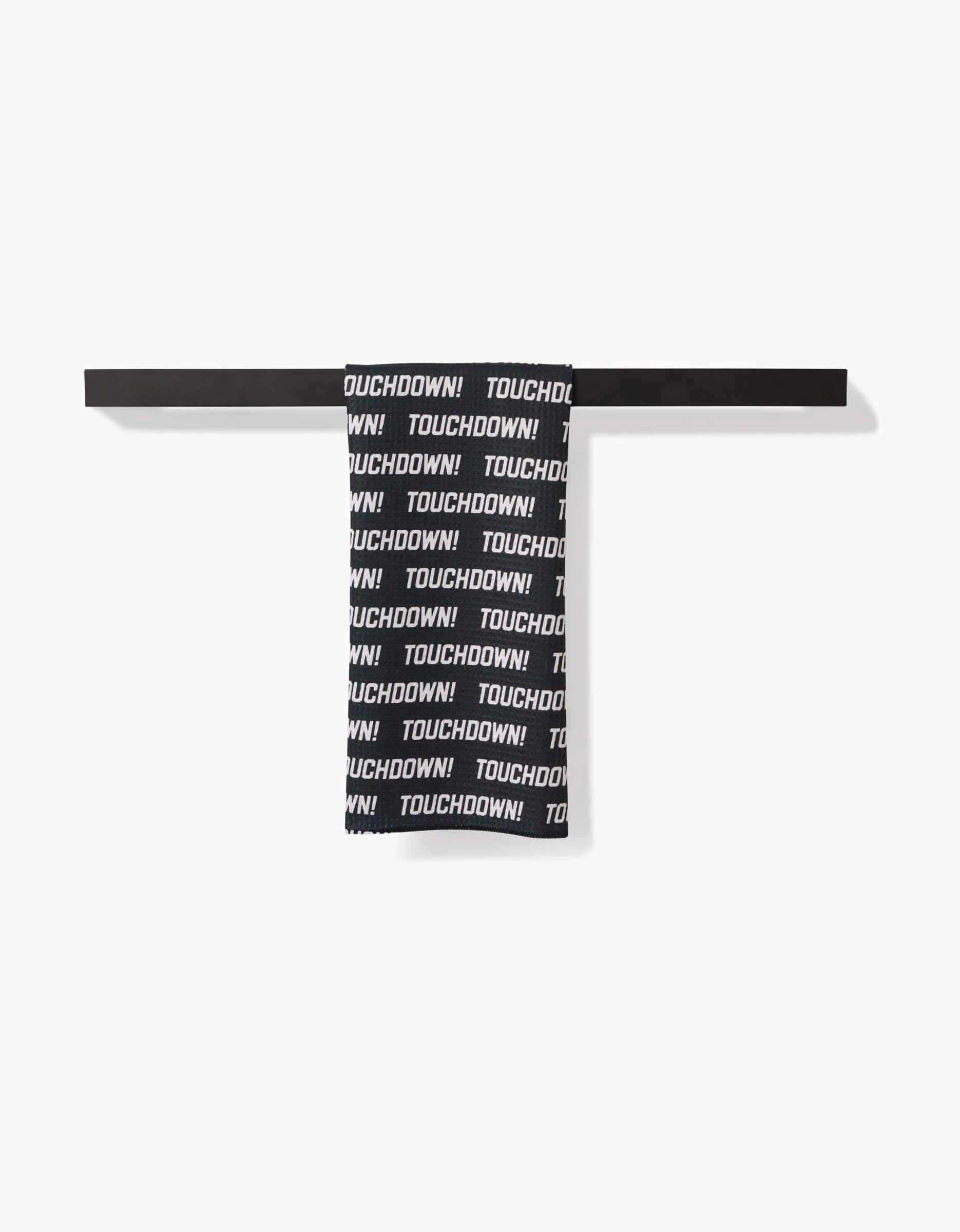 The GEOMETRY Touchdown tea towel, made from recycled materials, features bold black and white TOUCHDOWN! text and hangs neatly on a horizontal black rack against a white background.