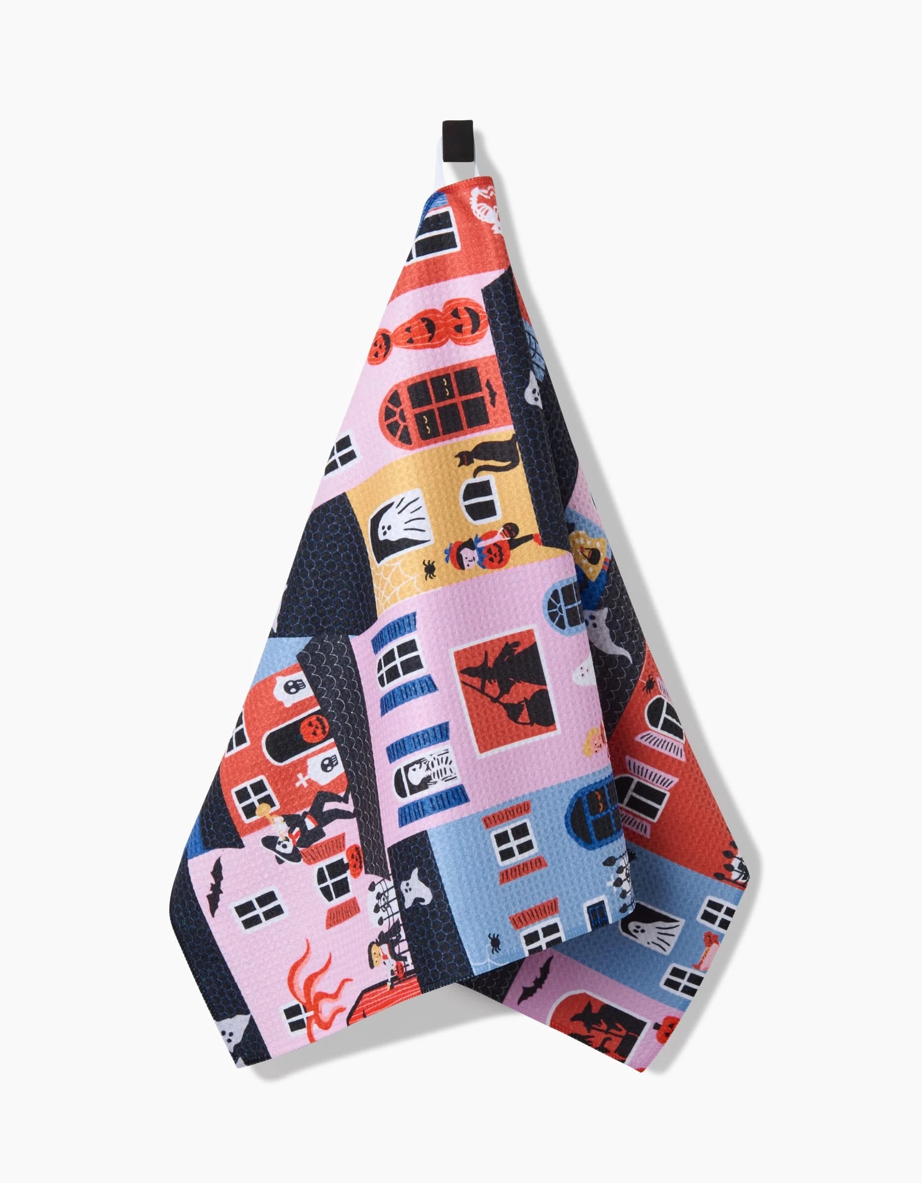 The Trick Or Treat Night kitchen towel by Ceyda Alasar is eco-friendly and features a vibrant pattern of houses, ghosts, pumpkins, and bats with a black hanging loop—all set against a white background.