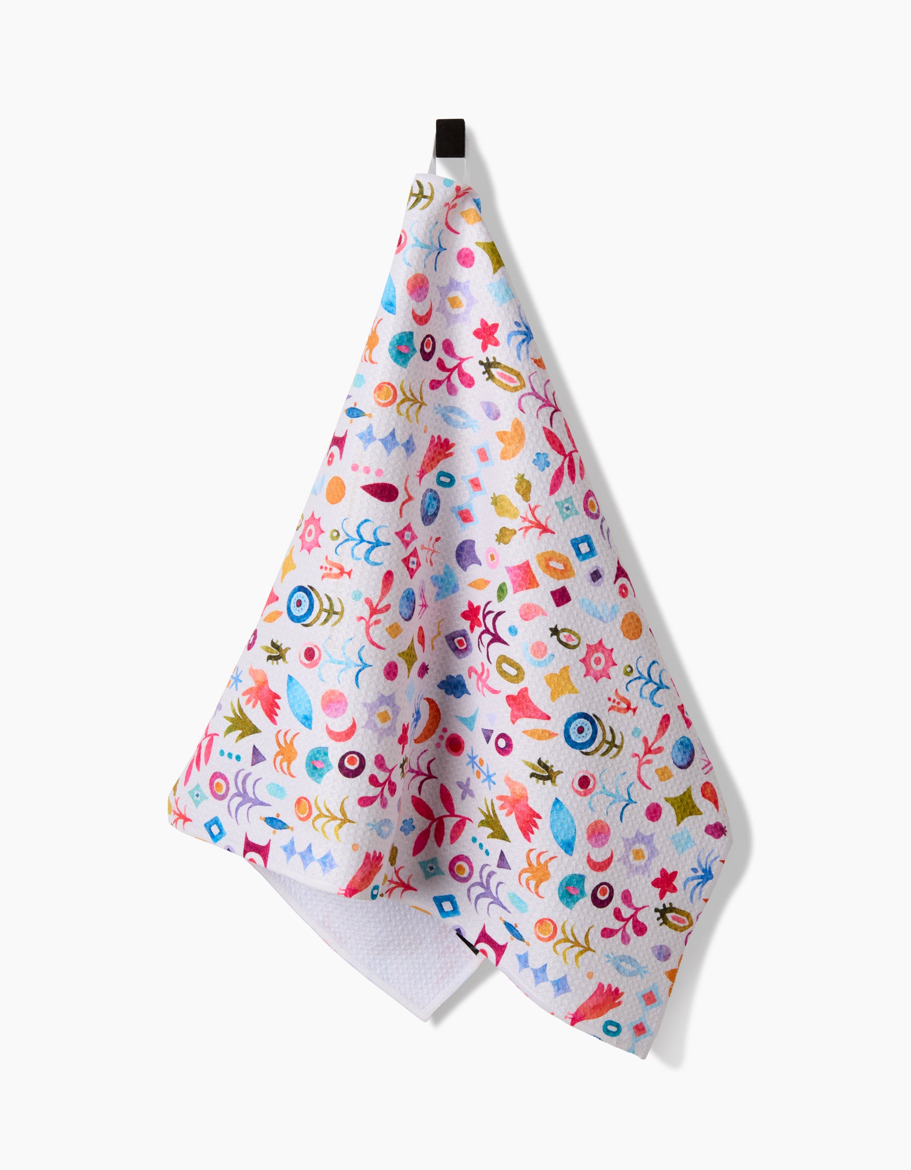 The Tula Tea Towel by Maayan Alper-Swan features a playful pattern of abstract shapes in blue, pink, yellow, orange, and red on white Geoweave fabric. It hangs on a black hook against a white background.