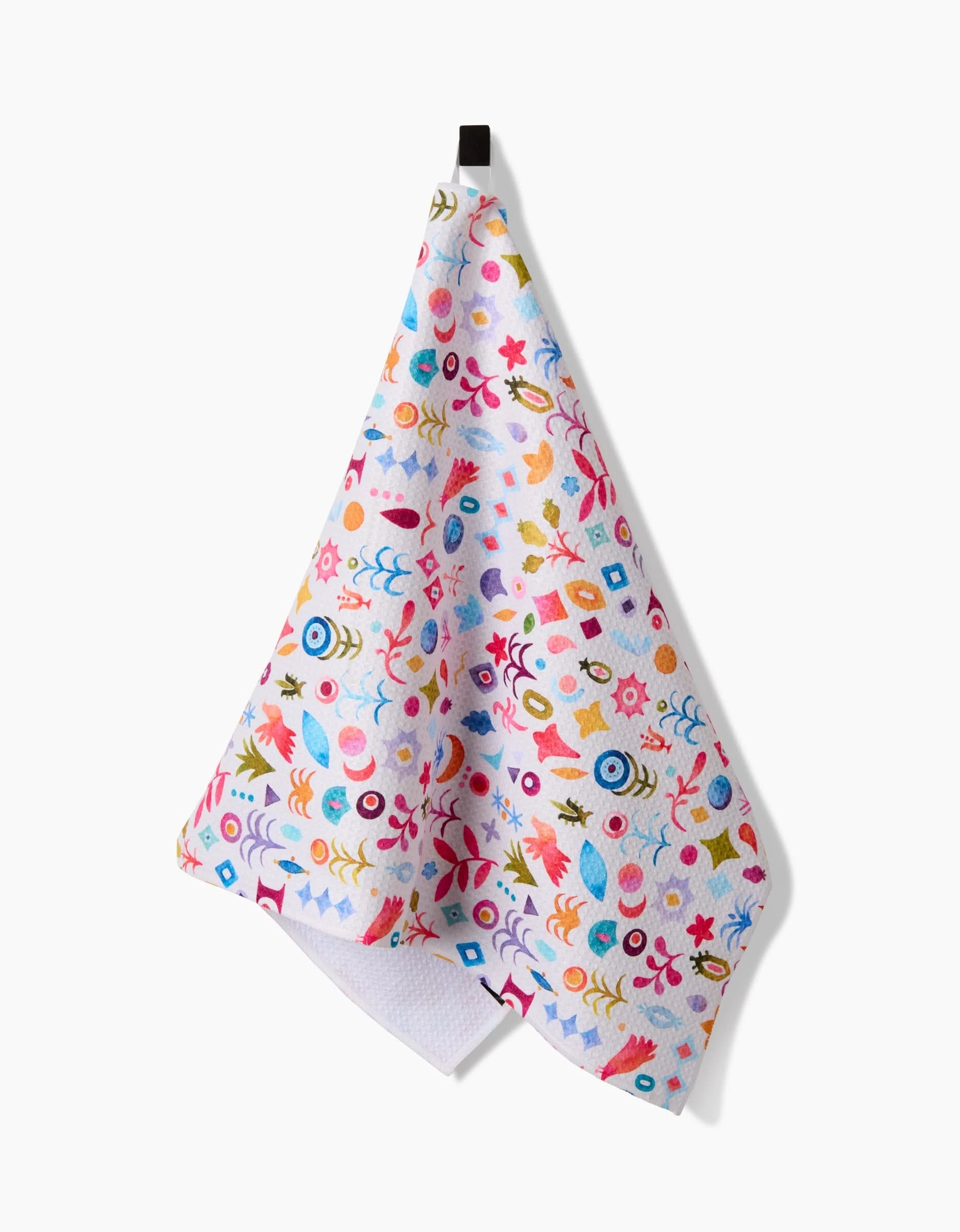The Tula Tea Towel by Maayan Alper-Swan features a playful pattern of abstract shapes in blue, pink, yellow, orange, and red on white Geoweave fabric. It hangs on a black hook against a white background.