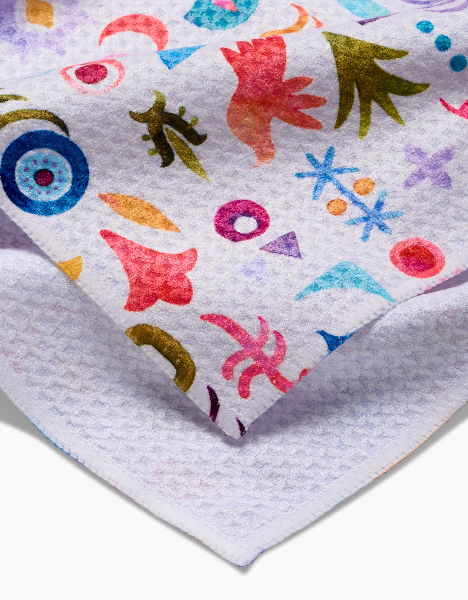 A close-up of the soft, textured white Geoweave fabric from Maayan Alper-Swan’s Tula tea towel, featuring colorful abstract birds, leaves, circles, and shapes in blue, pink, orange, and purple. The eco-friendly fabric is slightly folded.