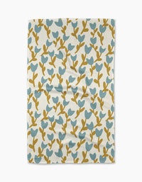 The Tulip Garden tea towel by Lizzie Clark is a rectangular, white towel made from recycled materials, featuring blue and mustard yellow stylized leaf and branch patterns. Displayed flat with slight wrinkles.