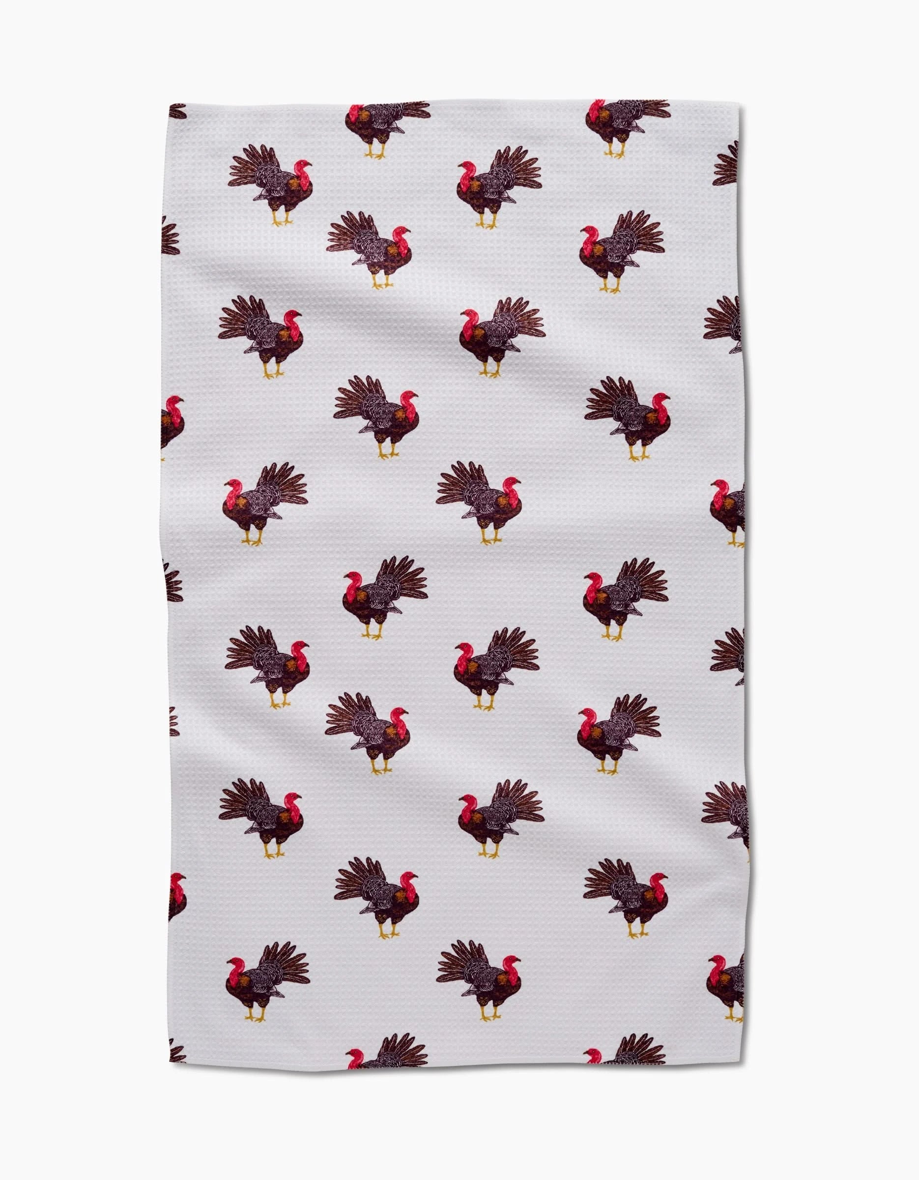 The Turkey Trot tea towel by Vaishali Design Studio features a white textured fabric with a neat, repeating pattern of illustrated turkeys—brown feathers, red wattles, and yellow legs—arranged in rows.