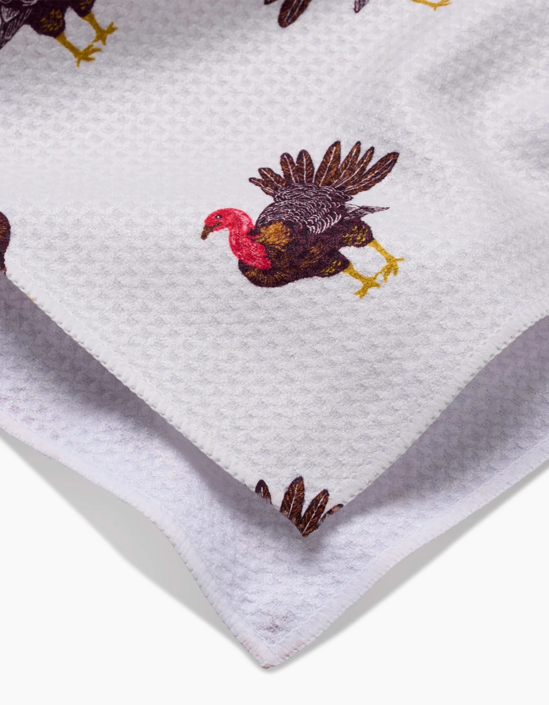 A close-up of the Vaishali Design Studio Turkey Trot tea towel, featuring a white textured fabric with a repeating pattern of turkeys with brown feathers, yellow legs, and red heads.