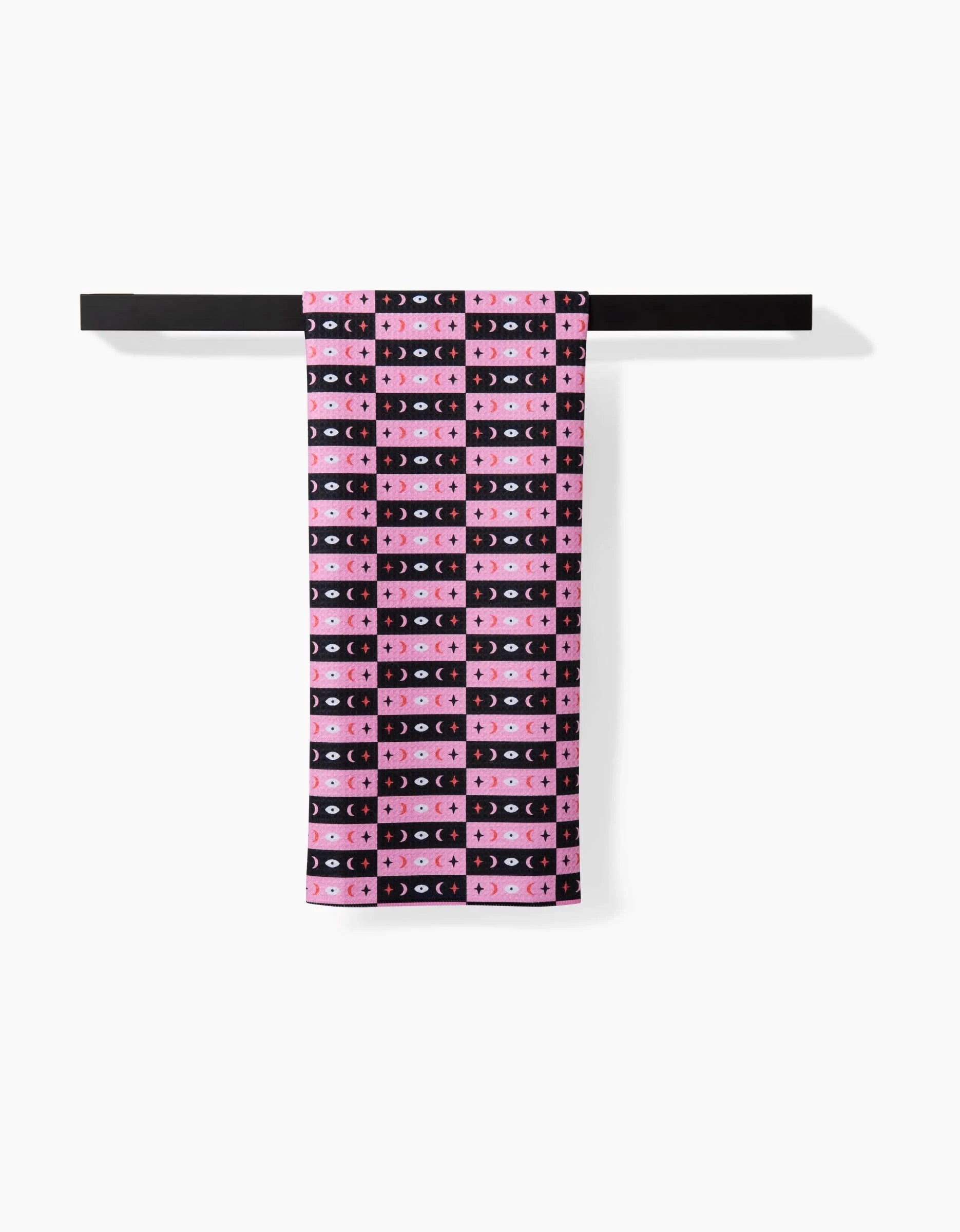The Laura Supnik Twinkling Eye tea towel, quick-drying and patterned with pink, black, and blue grids plus crescent moons and stars, hangs neatly folded over a black bar against a white background.