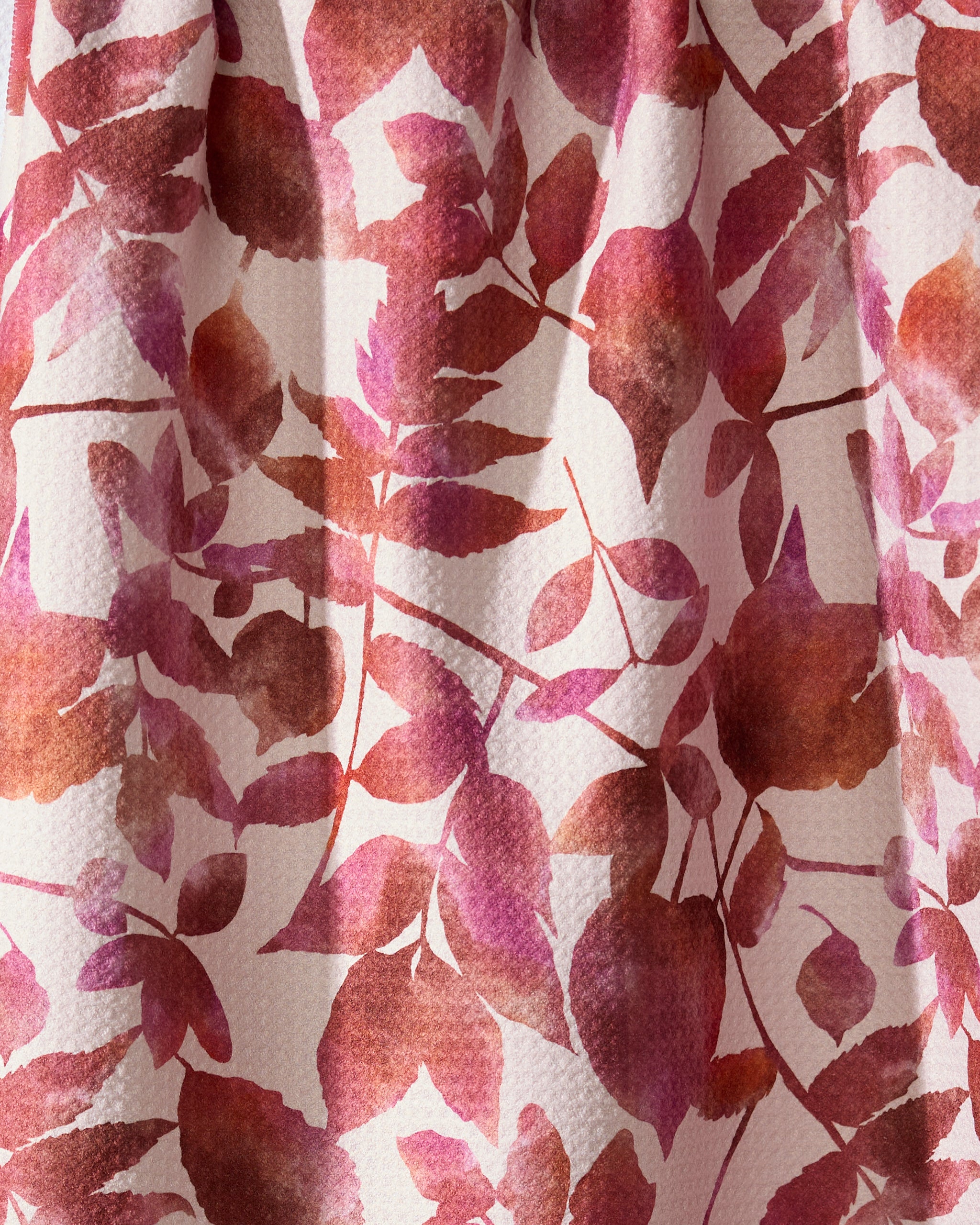 A close-up of Geometry’s Watercolor Dreaming tea towel, made from recycled materials, shows its pink, red, and brown leaf and flower pattern on a light background with a charmingly textured fabric.