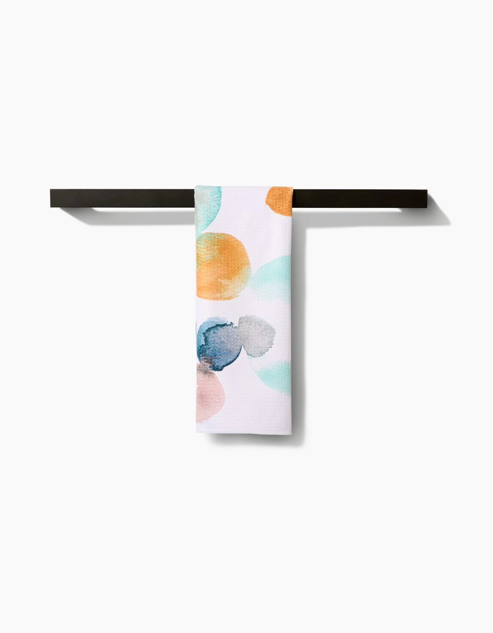 The Watercolor Rocks hand towel by Taryn Brennan, featuring vibrant abstract circles in orange, blue, teal, and peach on recycled fabric, hangs neatly on a dark towel bar against a white backdrop.