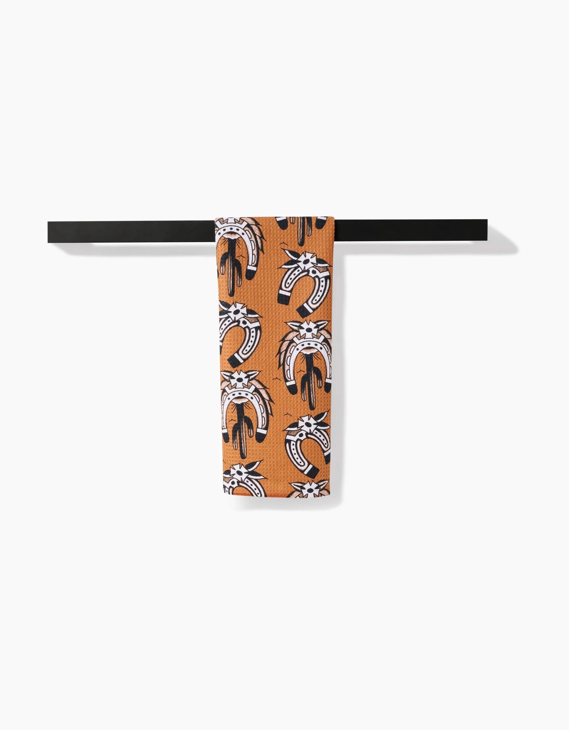 A Western Horseshoes tea towel by Cole Strem, featuring a repeating abstract animal face pattern, hangs neatly over a black bar. Its ultra-absorbent Geoweave fabric stands out against the crisp white background.
