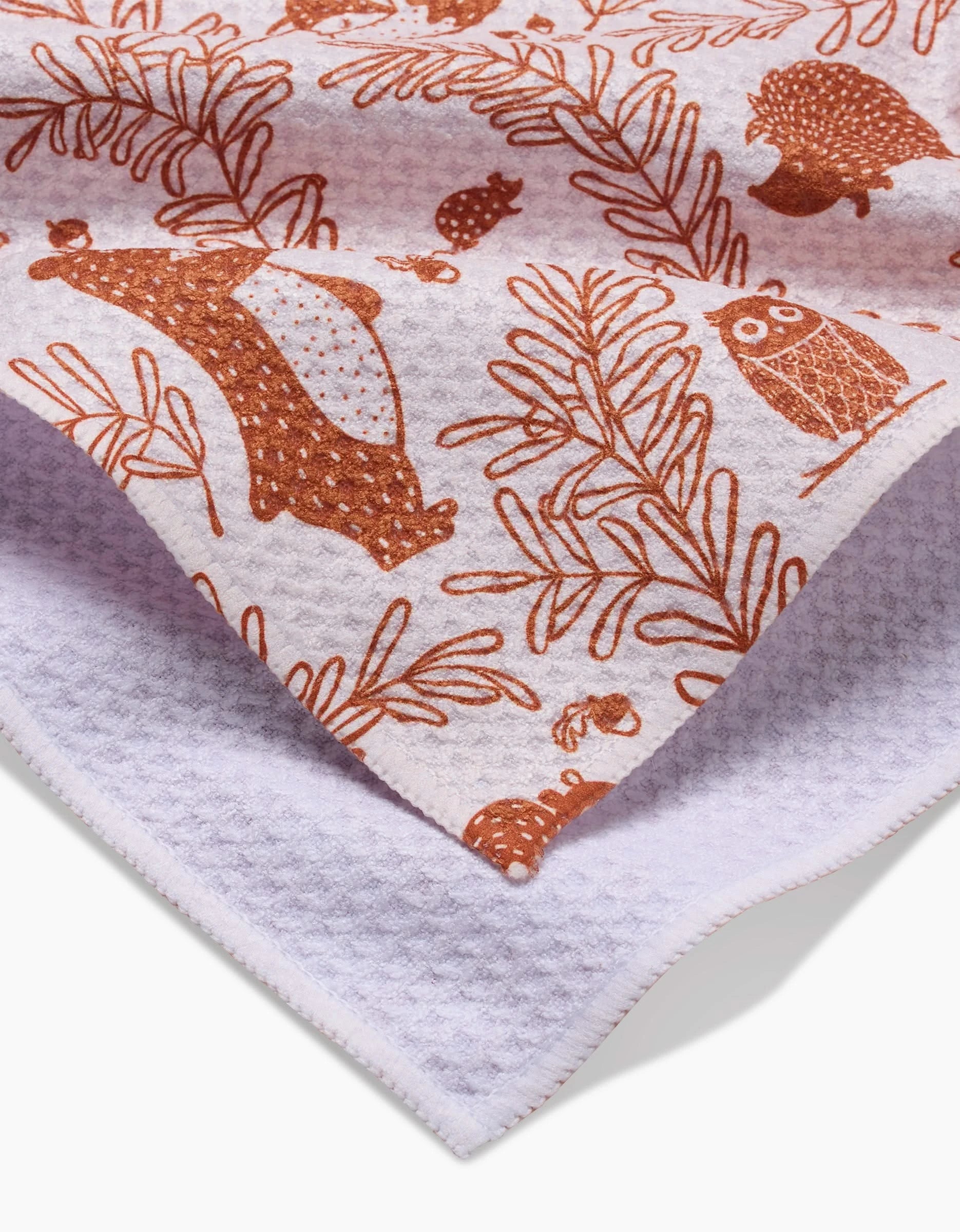 A close-up of the Sara Mari Whimsical Woodland ultra-absorbent bar towel, featuring brown bear, owl, hedgehog, and leaf illustrations on white textured fabric. The folded towel displays its patterned front and plain back.