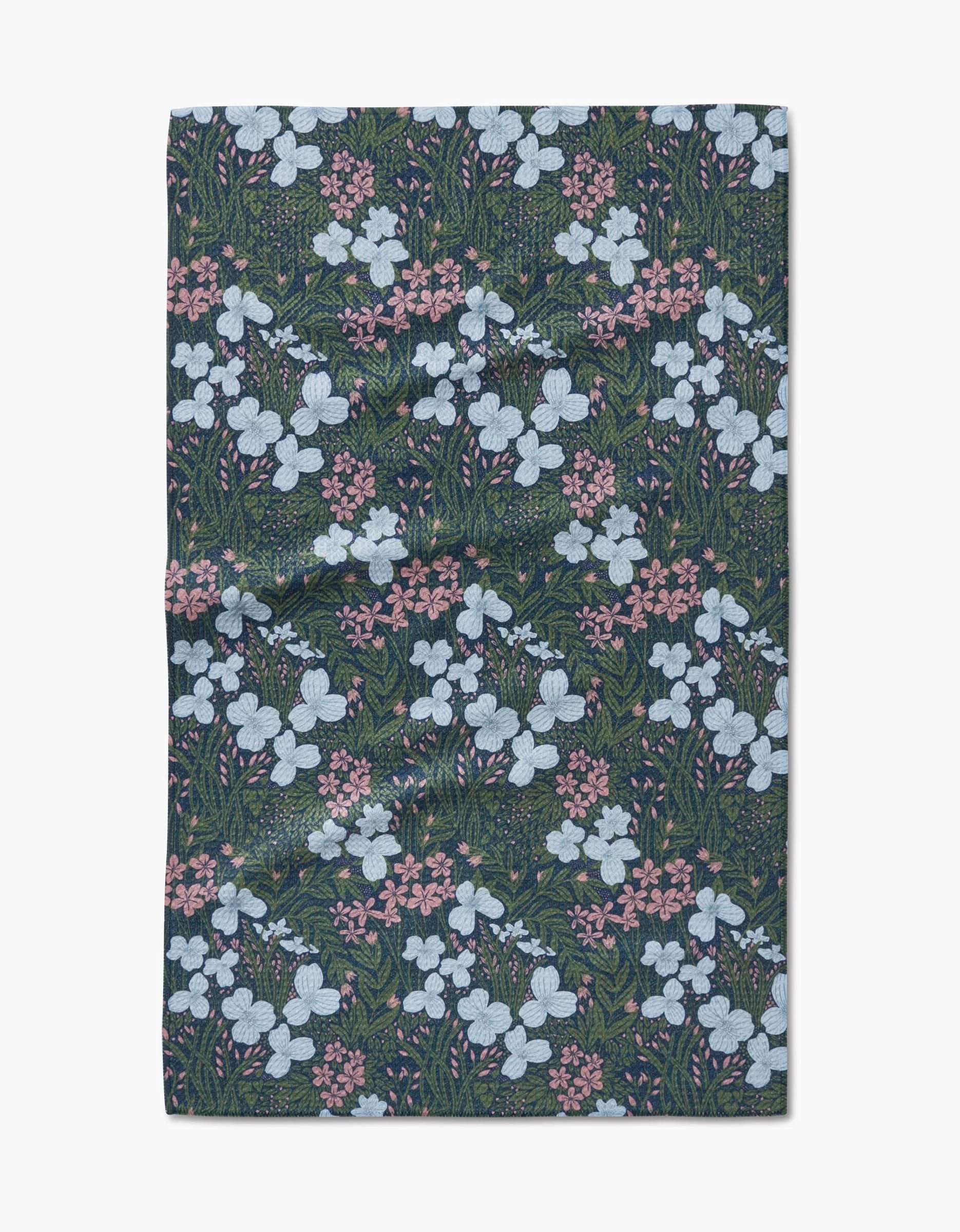 The White And Green Meadow Tea Towel by Katie OShea is crafted from Geoweave fabric and features a repeating pattern of white and pink flowers with green foliage on a dark background, shown flat on a white surface.