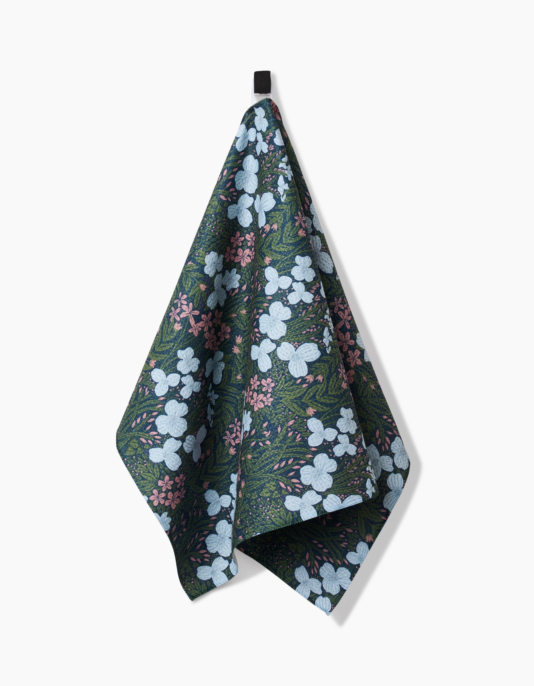 The White And Green Meadow tea towel by Katie OShea is made from recycled materials and showcases a botanical print of white and pink flowers with green leaves, hanging from a black loop against a plain white background.