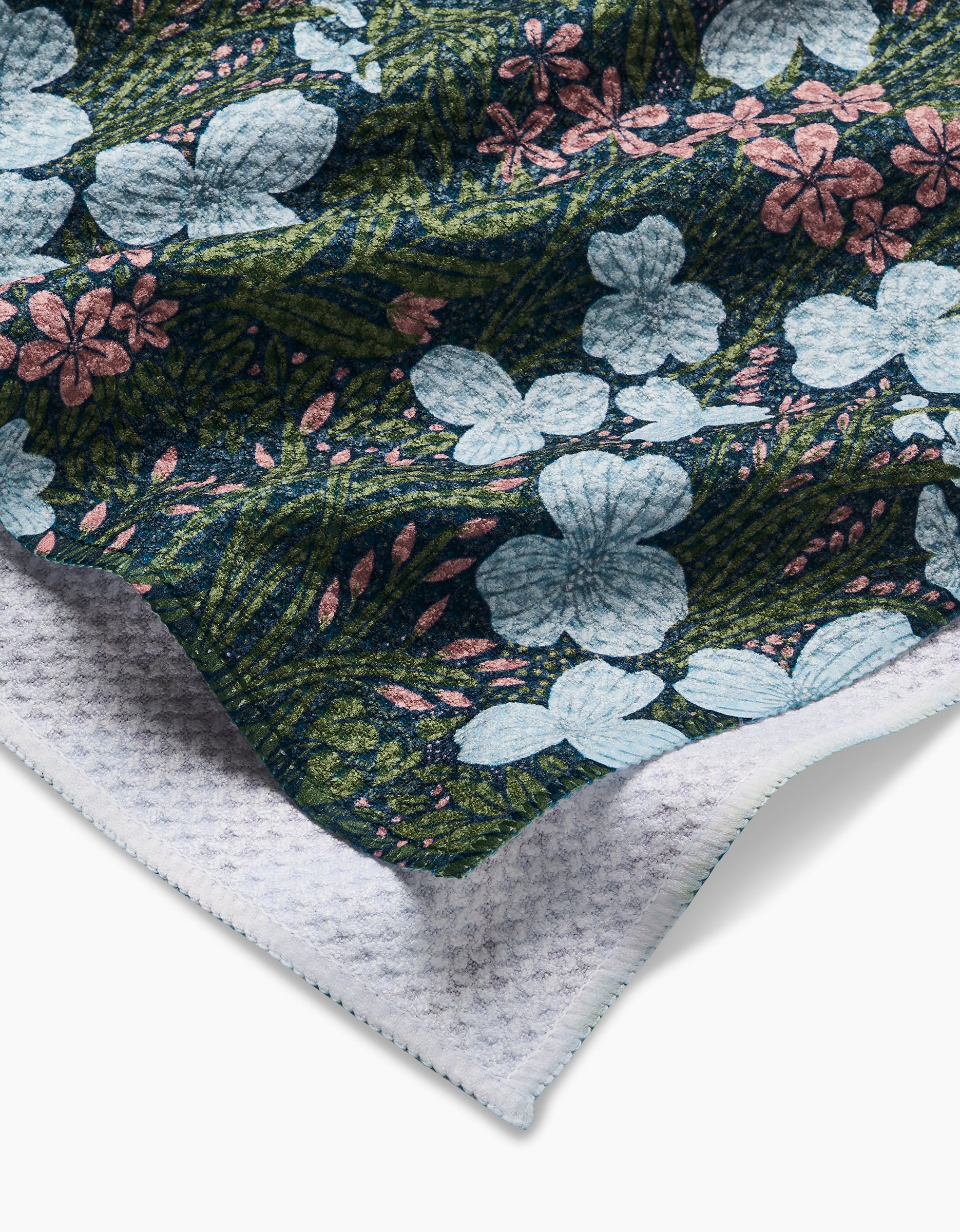 A close-up of Katie OSheas White And Green Meadow tea towel, crafted from recycled materials, showing large light blue and small pink flowers on a dark green leafy background with a folded white waffle-patterned corner.