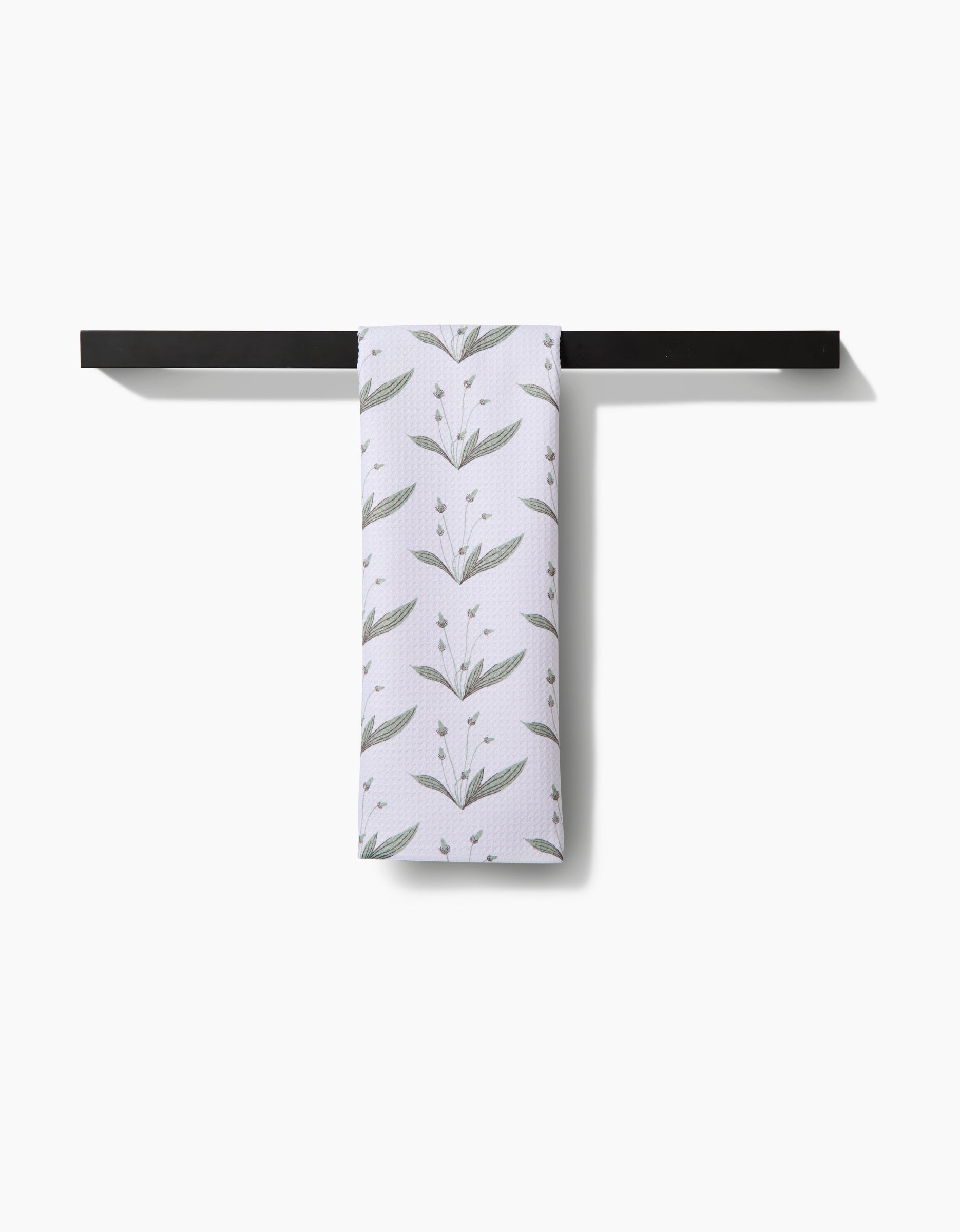 The Linda Fox Wild Leaves Green tea towel, featuring a green leafy branch pattern on white Geoweave fabric, hangs neatly on a black towel bar against a plain white background, offering style and superior absorbency.
