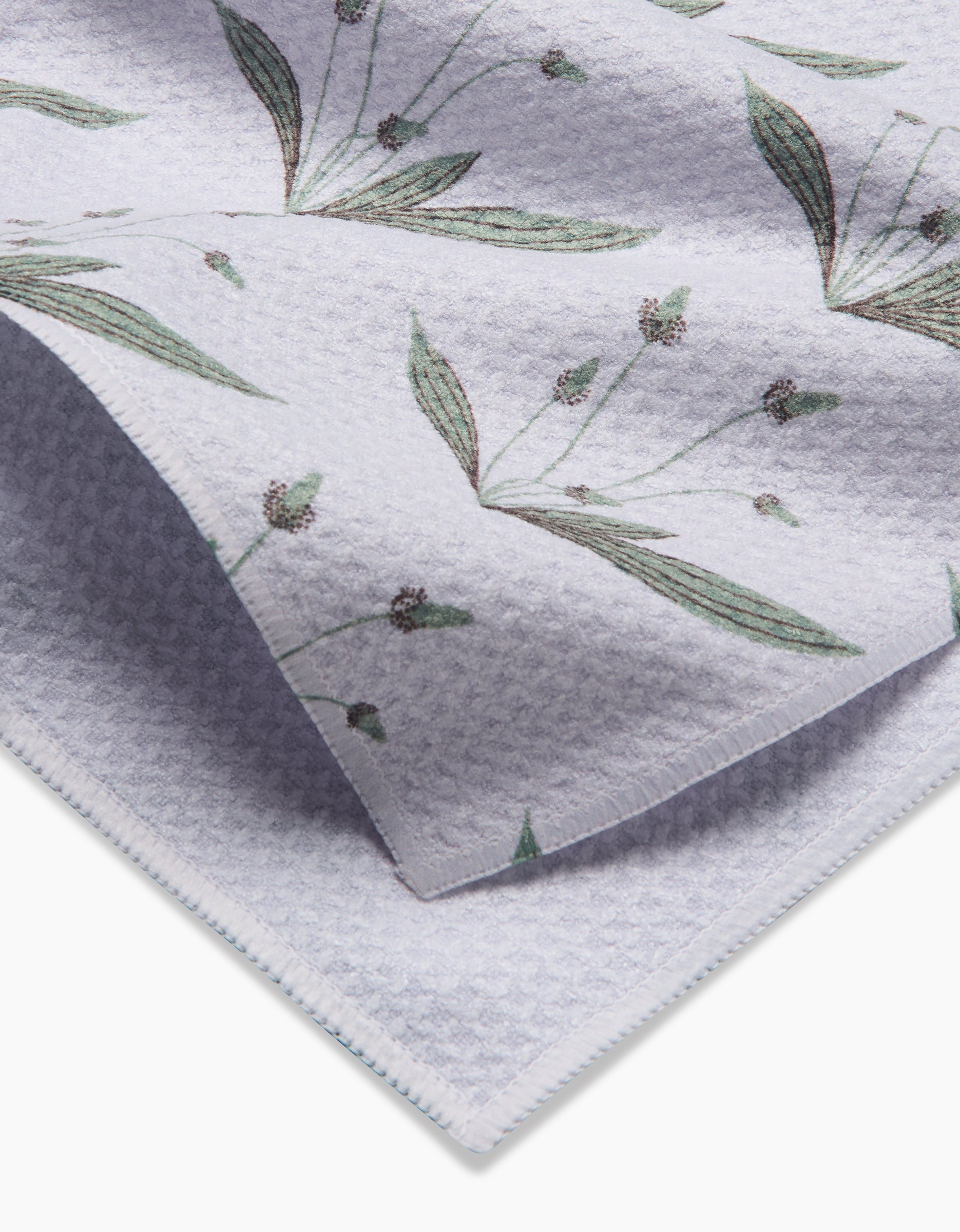 A close-up of the Linda Fox Wild Leaves Green tea towel shows its textured, light fabric with a green leaf and small bud print. The photo highlights folded Geoweave material, displaying both front and back sides.