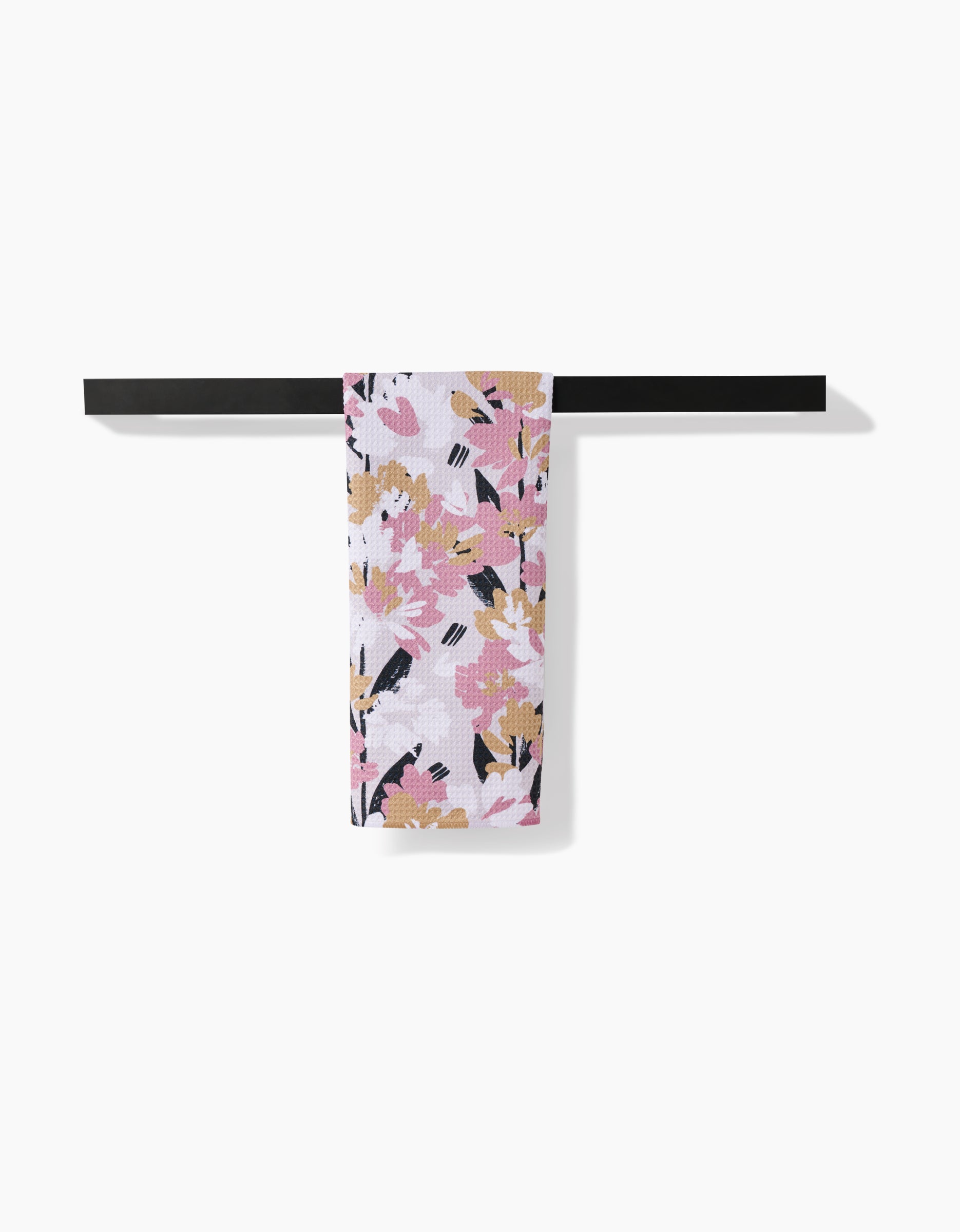 The Toboggan Avenue Wilderness Floral tea towel, featuring pink, gold, white, and black floral patterns and made from recycled materials, hangs neatly on a minimalist black towel bar against a plain white background.
