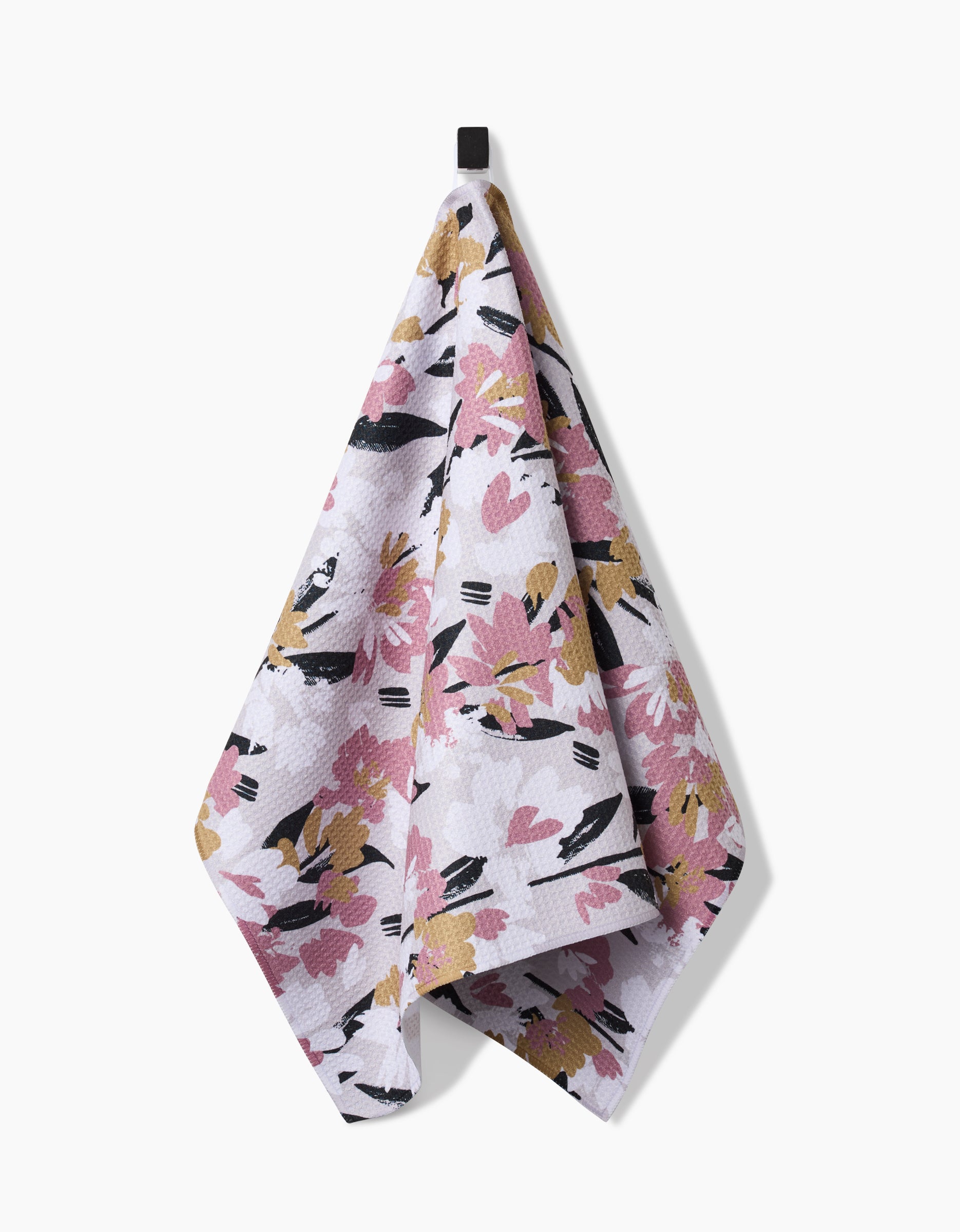 The Wilderness Floral tea towel by Toboggan Avenue hangs from a black hook, displaying pink, yellow, and black abstract floral designs on white Geoweave fabric.