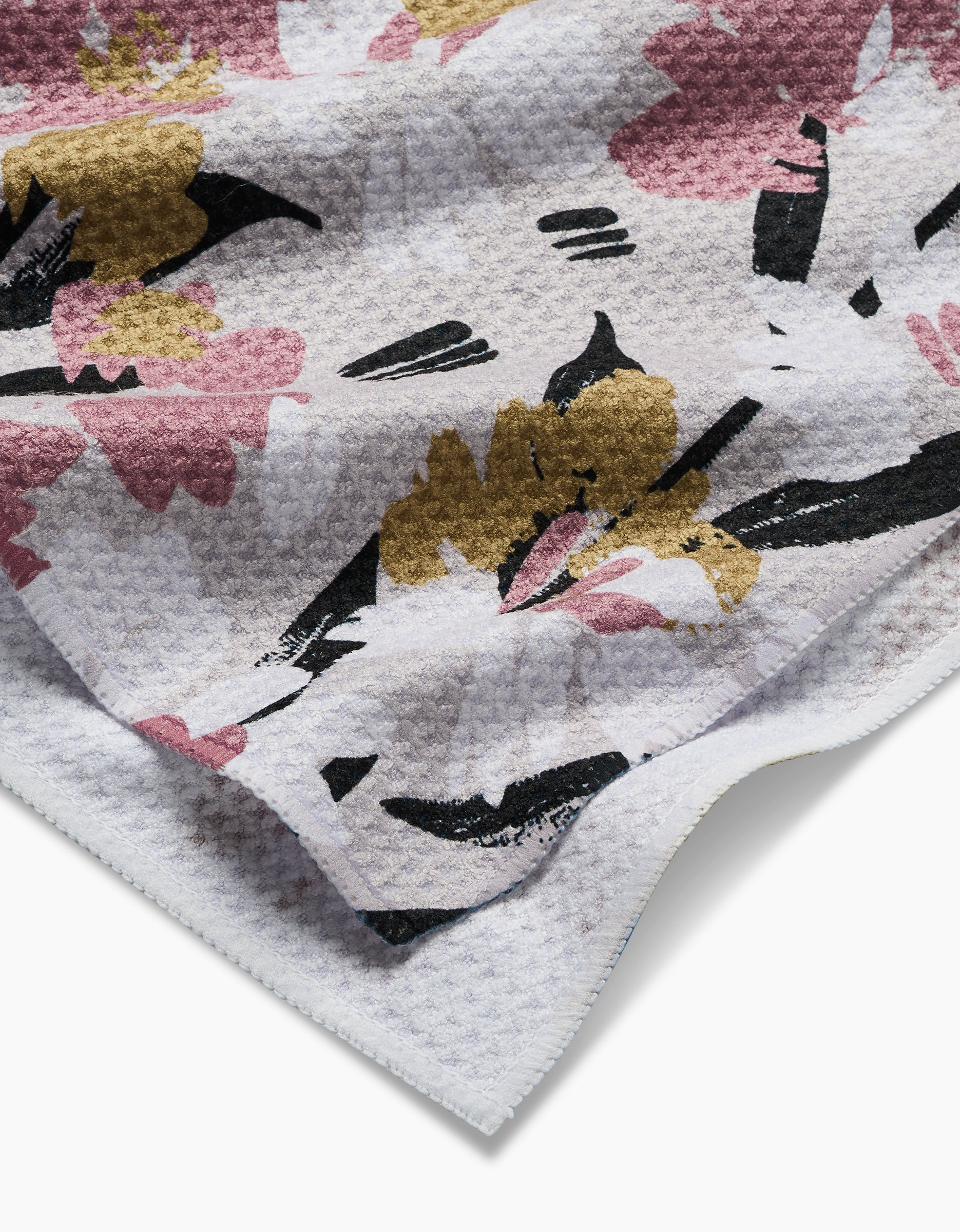 The Wilderness Floral tea towel by Toboggan Avenue features a textured white Geoweave fabric with a waffle pattern and a floral print in pink, mustard yellow, and black. The soft towel has slightly folded edges.