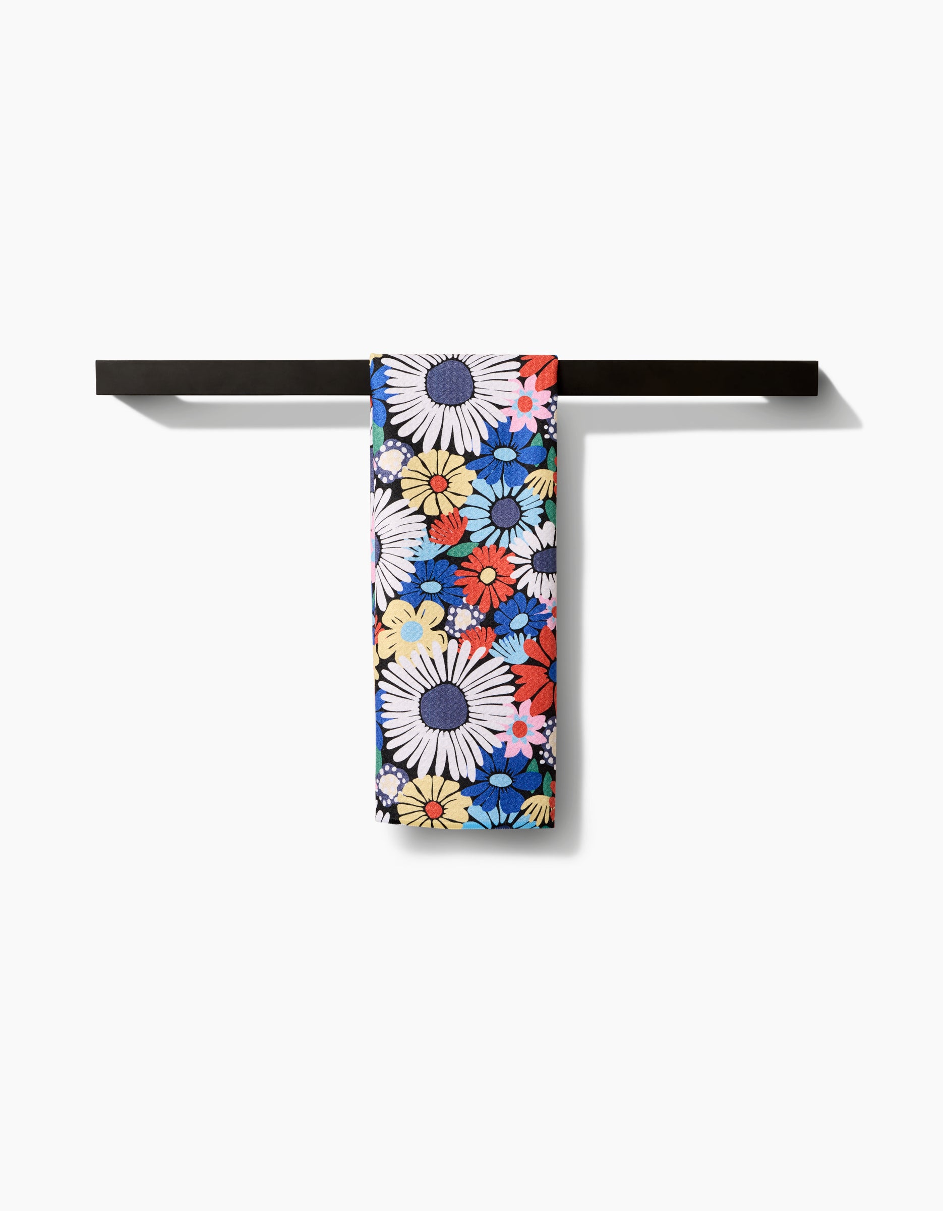 The Wildflowers Bouquet tea towel by Claire Iglesias, featuring large daisy designs in white, yellow, blue, red, and orange on recycled fabric, hangs on a black bar and stands out against a crisp white backdrop.