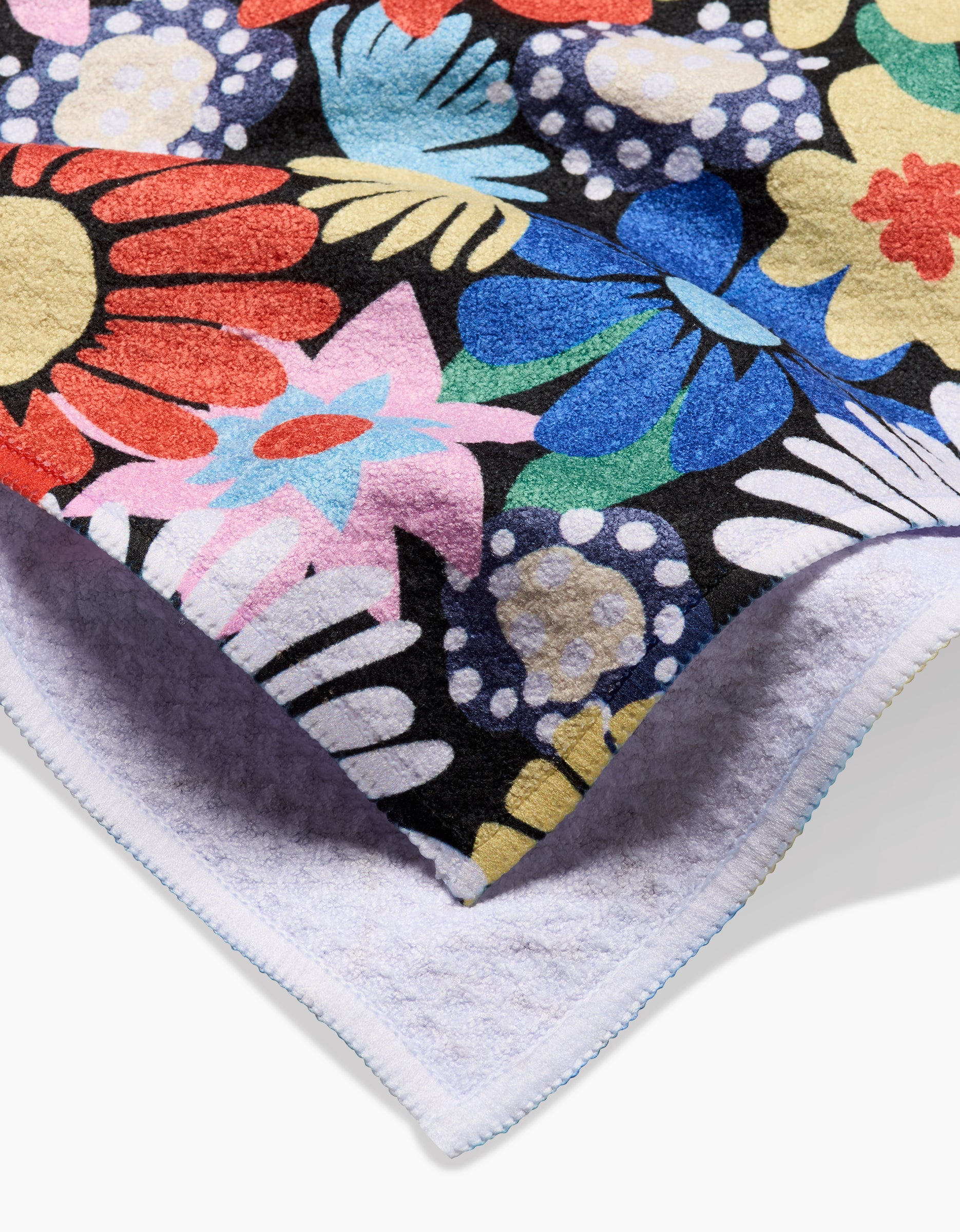 A close-up of Claire Iglesias’ Wildflowers Bouquet tea towel shows a bold, colorful floral pattern on black with red, blue, yellow, white, and pink blooms. The folded edge reveals the soft Geoweave™ fabric underside.