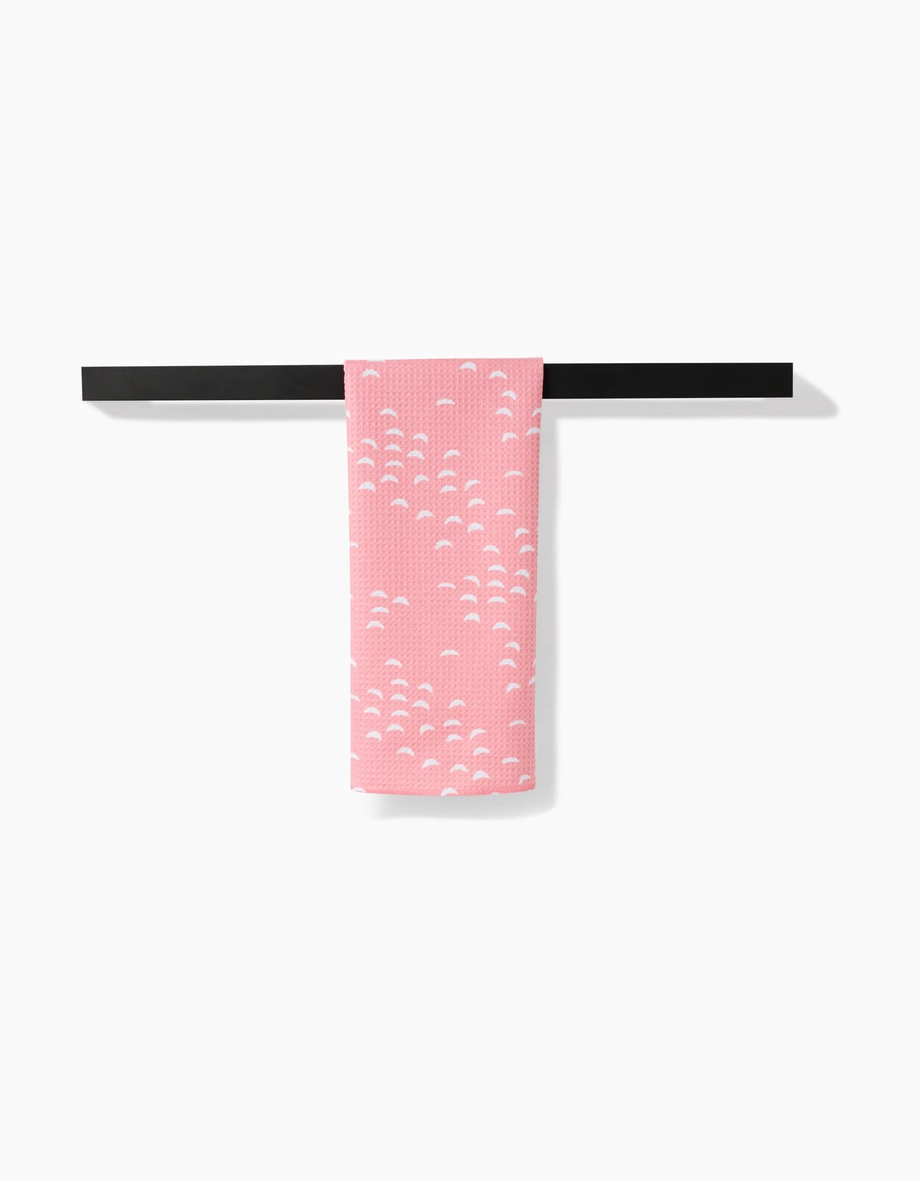 The Windswept tea towel by Jessica Swift, featuring a pink background with small white semi-circles, hangs neatly on a black bar against a plain white backdrop.