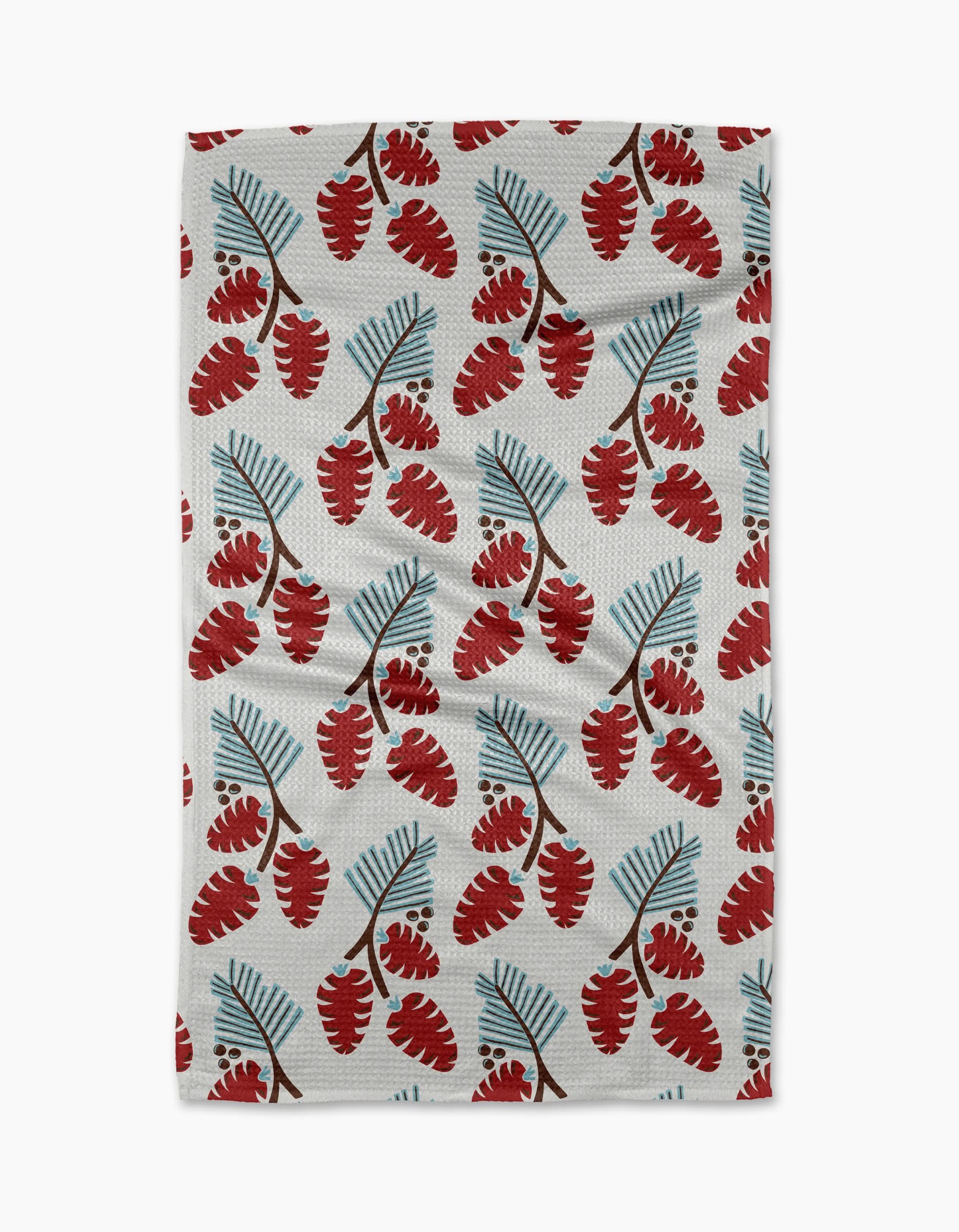The Winter Forage kitchen towel by Lizzie Clark is crafted from recycled white fabric and showcases a festive, nature-inspired design with red pinecones and blue-green pine needles in a repeating pattern.