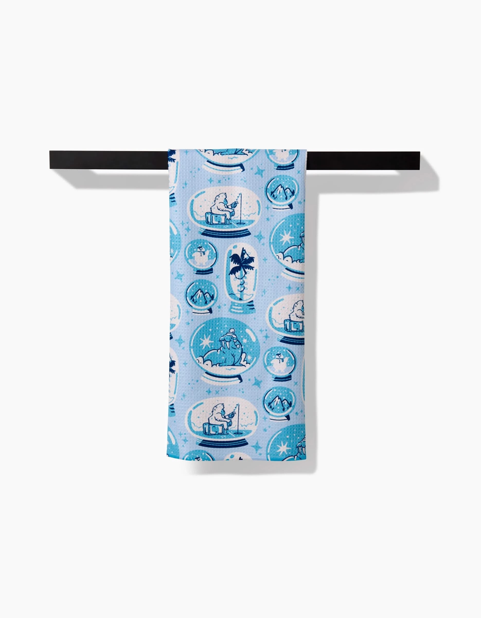 The Carly Berry Winter Snow Globe Collection tea towel, made from recycled materials, features light blue fabric with white and dark blue cartoon polar bears and snowy scenes, hanging on a black bar against a white background.