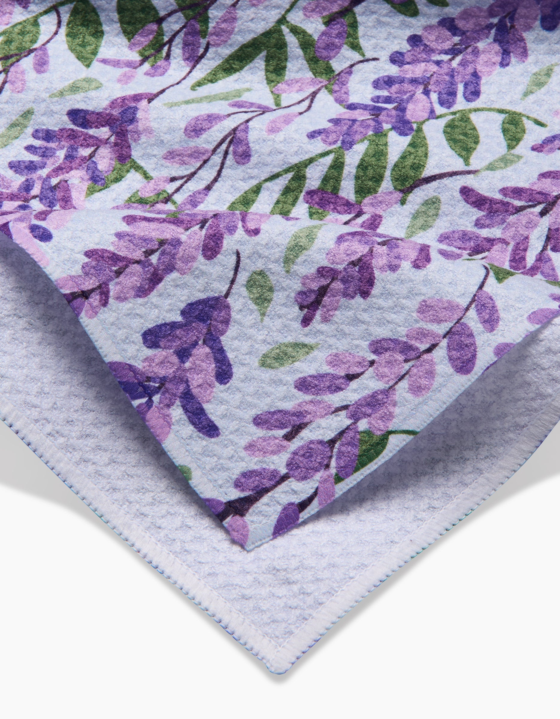 Close-up of Rebecca Bobkos Wisteria towels: the top towel is adorned with purple flowers and green leaves, while the bottom is plain and light. Both are ultra-absorbent for comfort and quick-drying convenience.