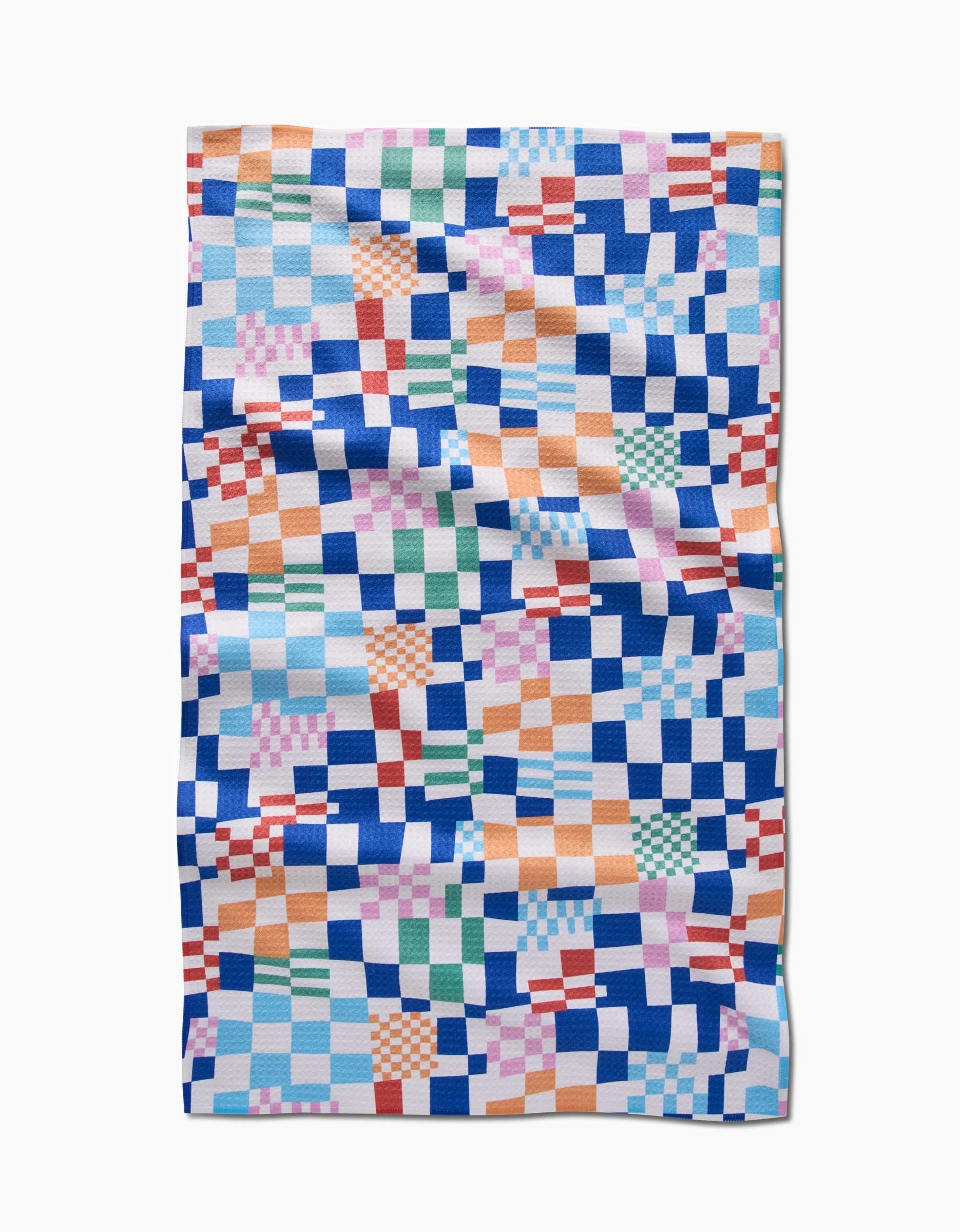 The Wonky Checkers tea towel by Claire Iglesias features a colorful, irregular checkerboard in blue, orange, green, pink, and white squares. Made from recycled materials, it’s quilted and shown flat with visible folds and creases.