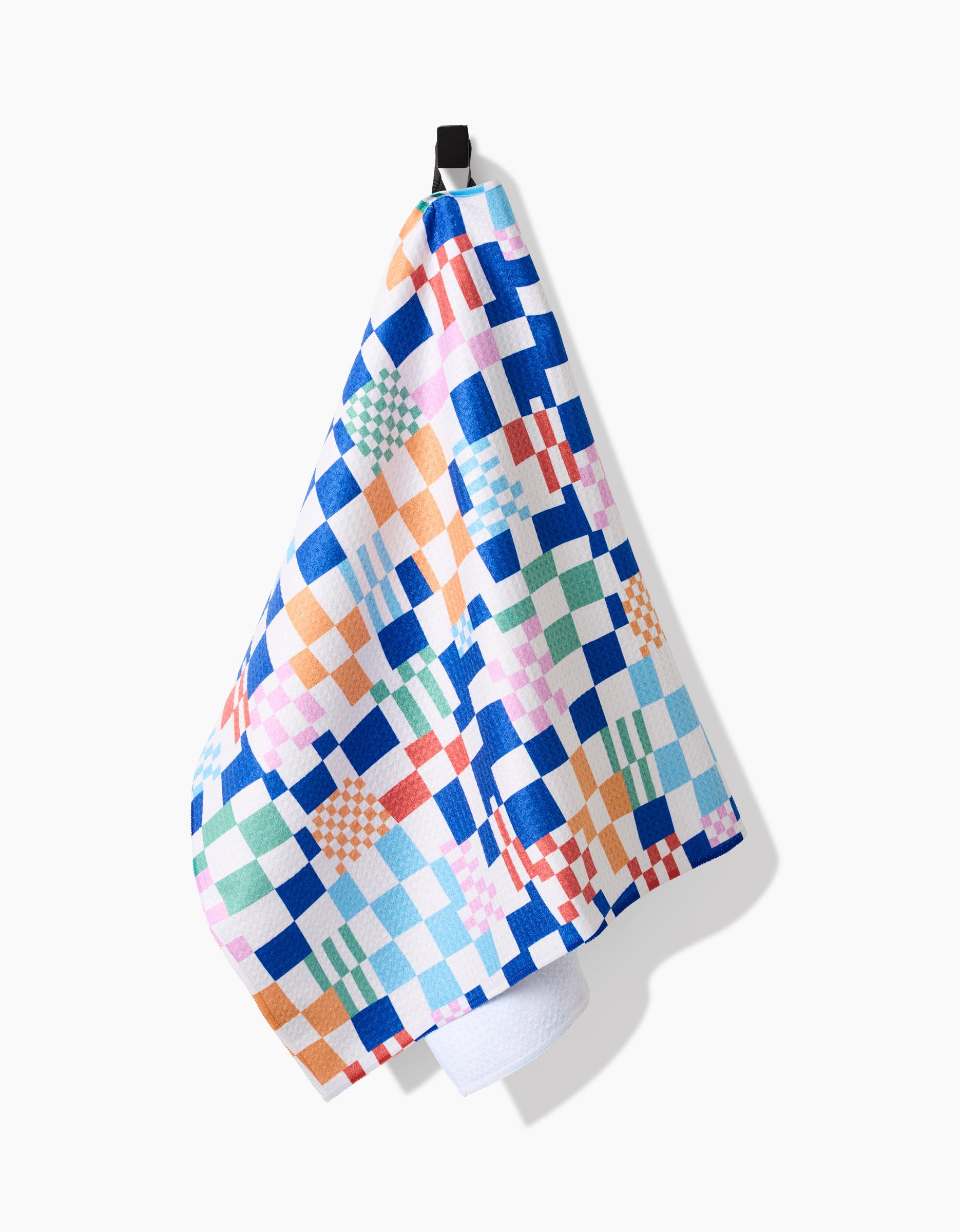 The Wonky Checkers tea towel by Claire Iglesias, made from recycled materials, displays blue, orange, green, pink, and white squares and hangs on a black hook against a white background.