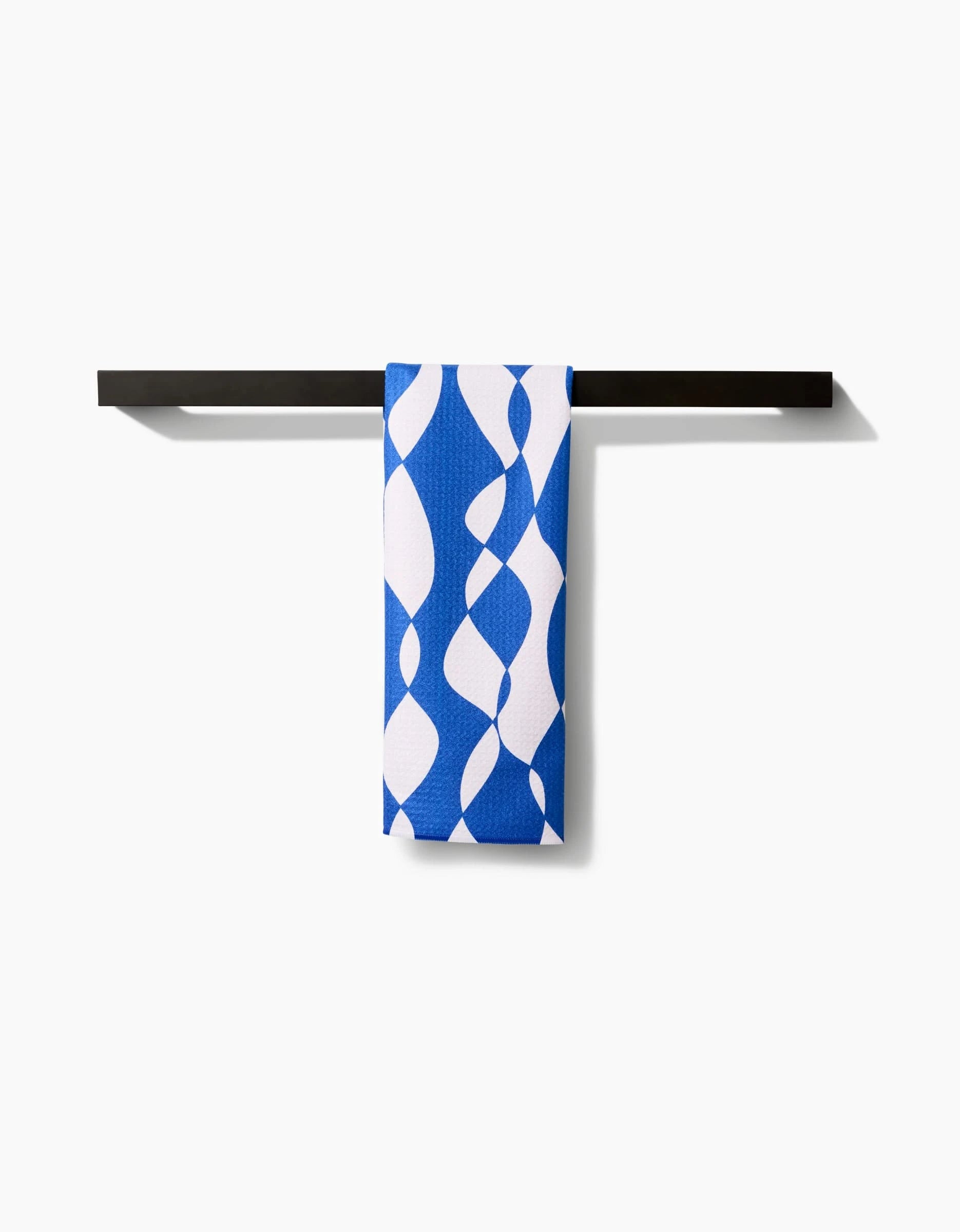 The Wonky Lines tea towel by Claire Iglesias, made from Geoweave fabric and featuring a blue and white abstract curvy design, hangs neatly on a simple black towel bar against a plain white background.