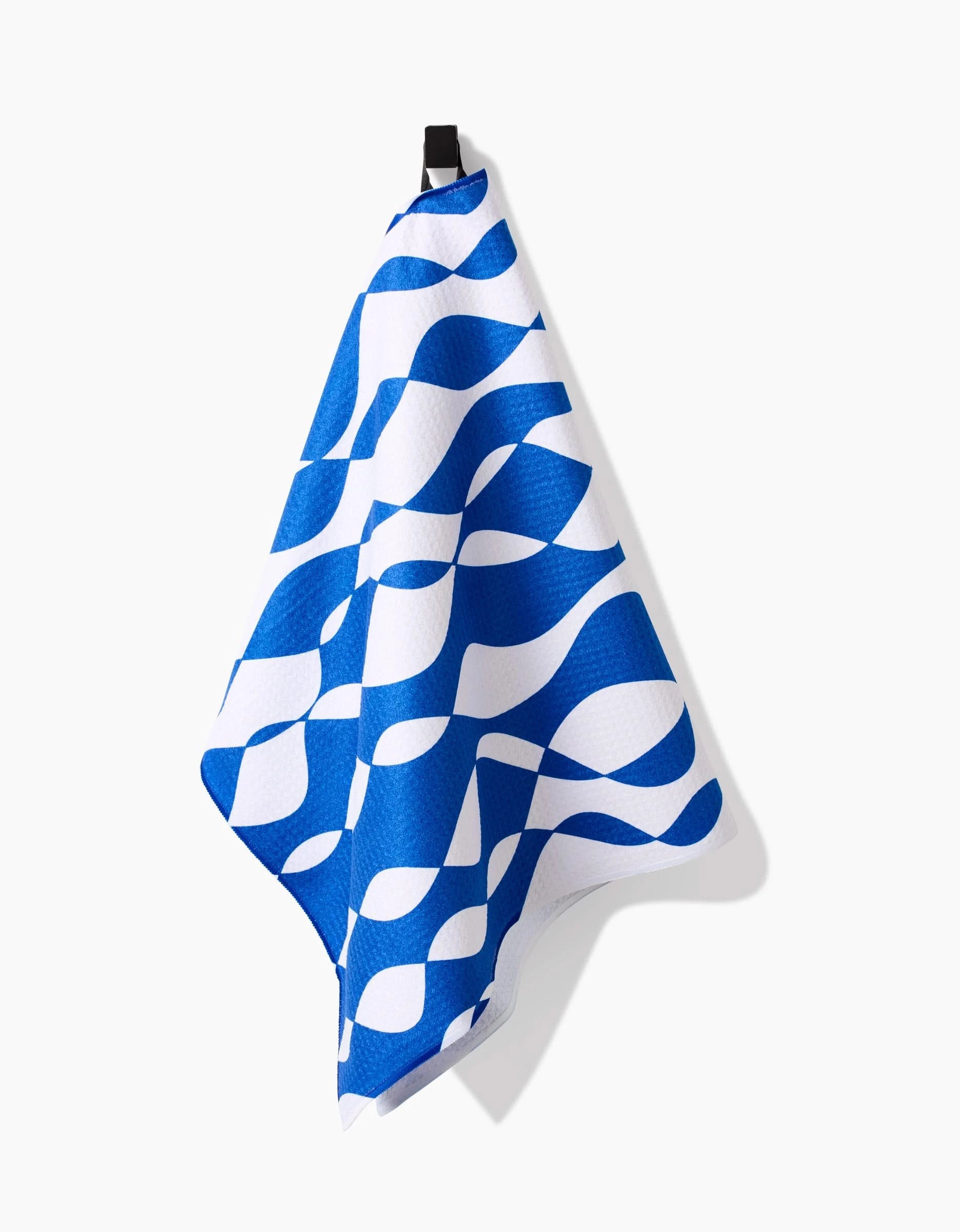 The Wonky Lines tea towel by Claire Iglesias, featuring abstract blue and white wave patterns and made from recycled materials, hangs on a black hook against a plain white background.